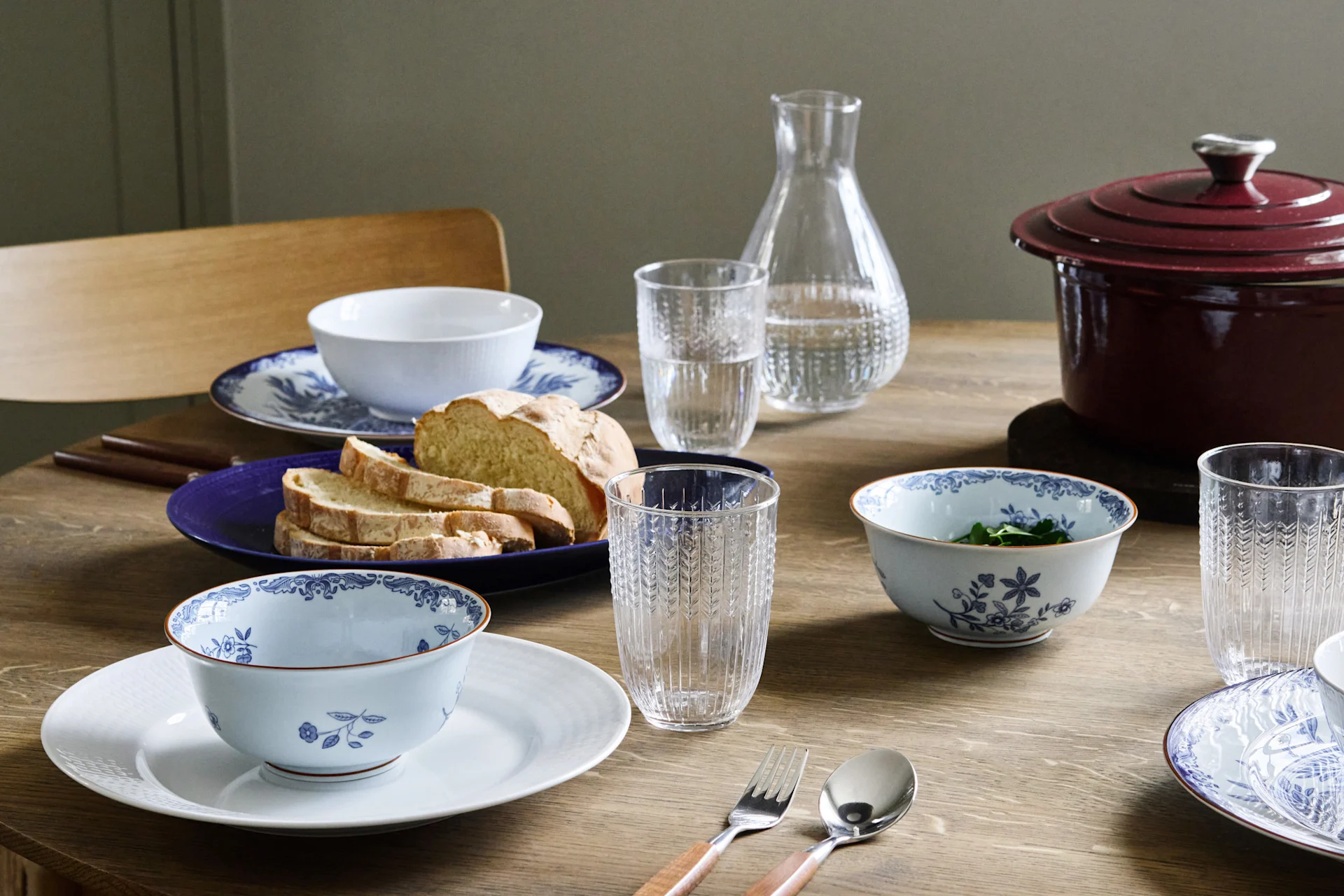 Ett träbord dukat med blå- och vitmönstrade skålar, tallrikar från Rörstrand, skivat bröd, vattenglas och en röd gryta med lock.