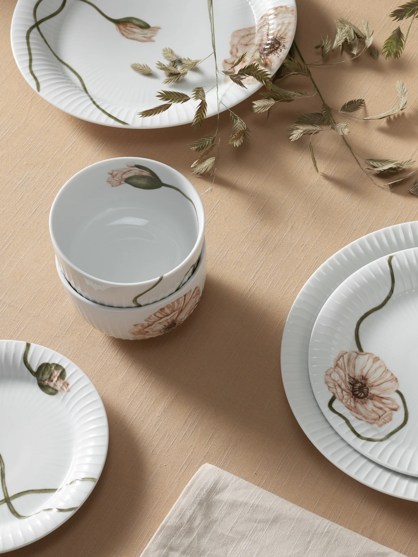Vita keramiska tallrikar och skålar med ljusorange vallmoblommor och räfflad textur på en ljusbrun duk med torkat gräs.