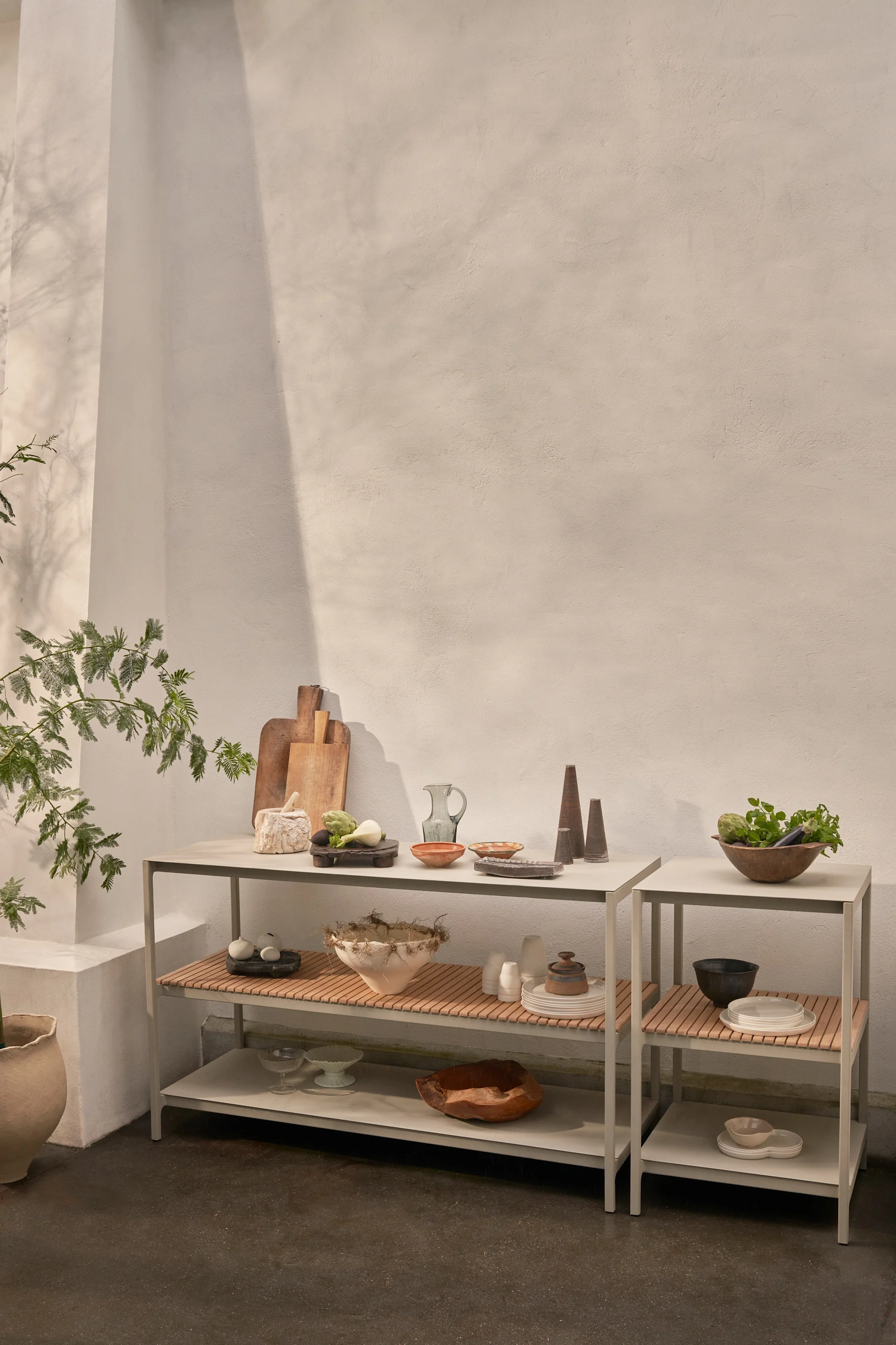 Två moderna grå hyllenheter med träribbor visar olika köksredskap, keramikskålar, färska grönsaker och dekorativa föremål mot en ljus vägg.