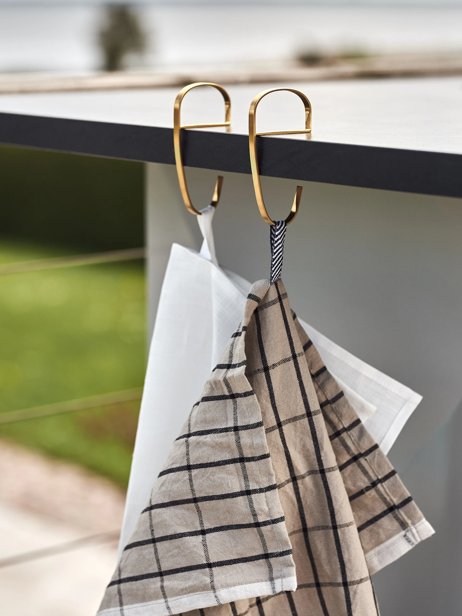 Två kökshanddukar, en vit och en beige rutig, hänger från gyllene S-krokar på en mörk bänk.