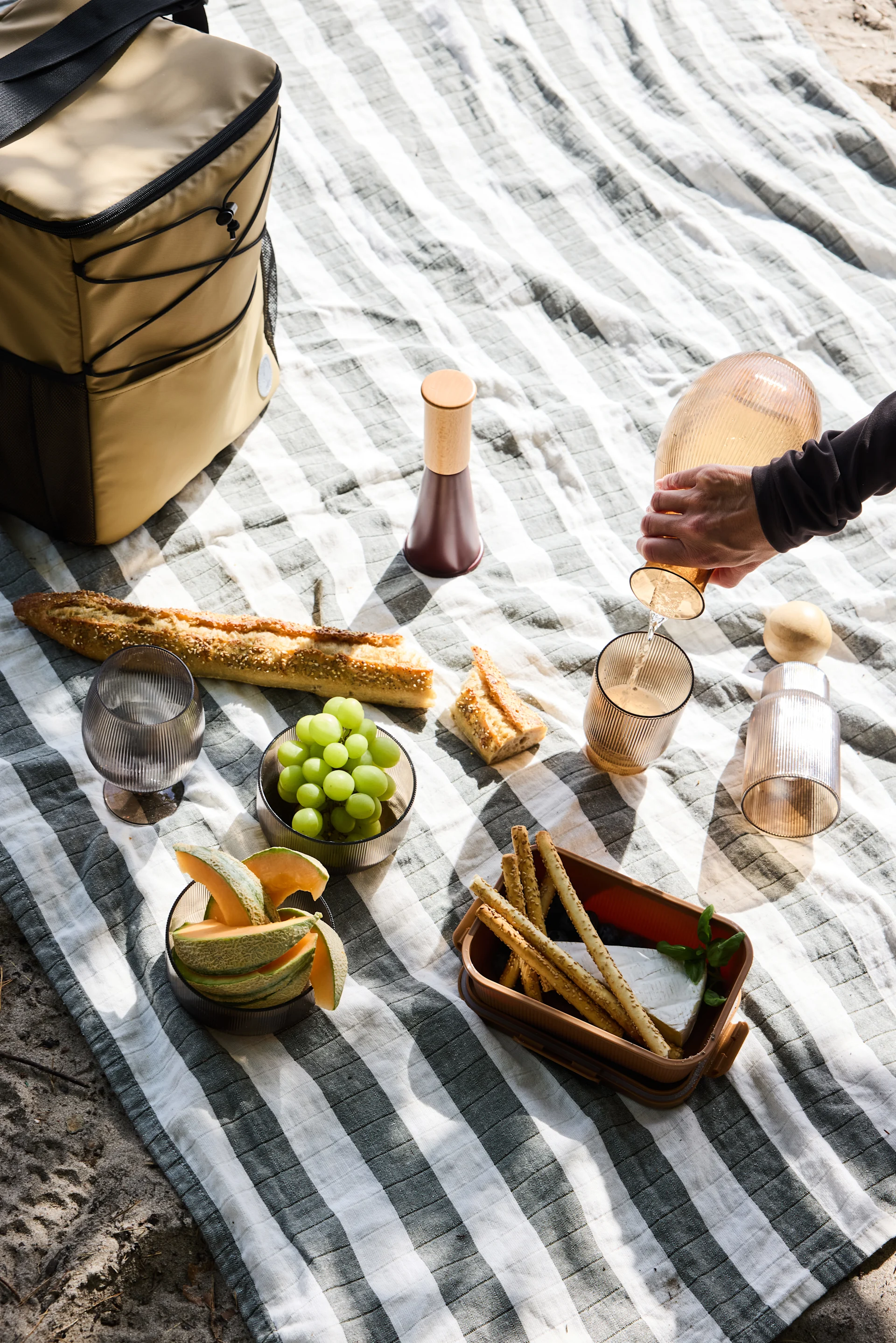 Översiktsbild av en picknick på en randig filt, med en person som häller från en räfflad karaff, baguette, frukt, ost och brödpinnar.
