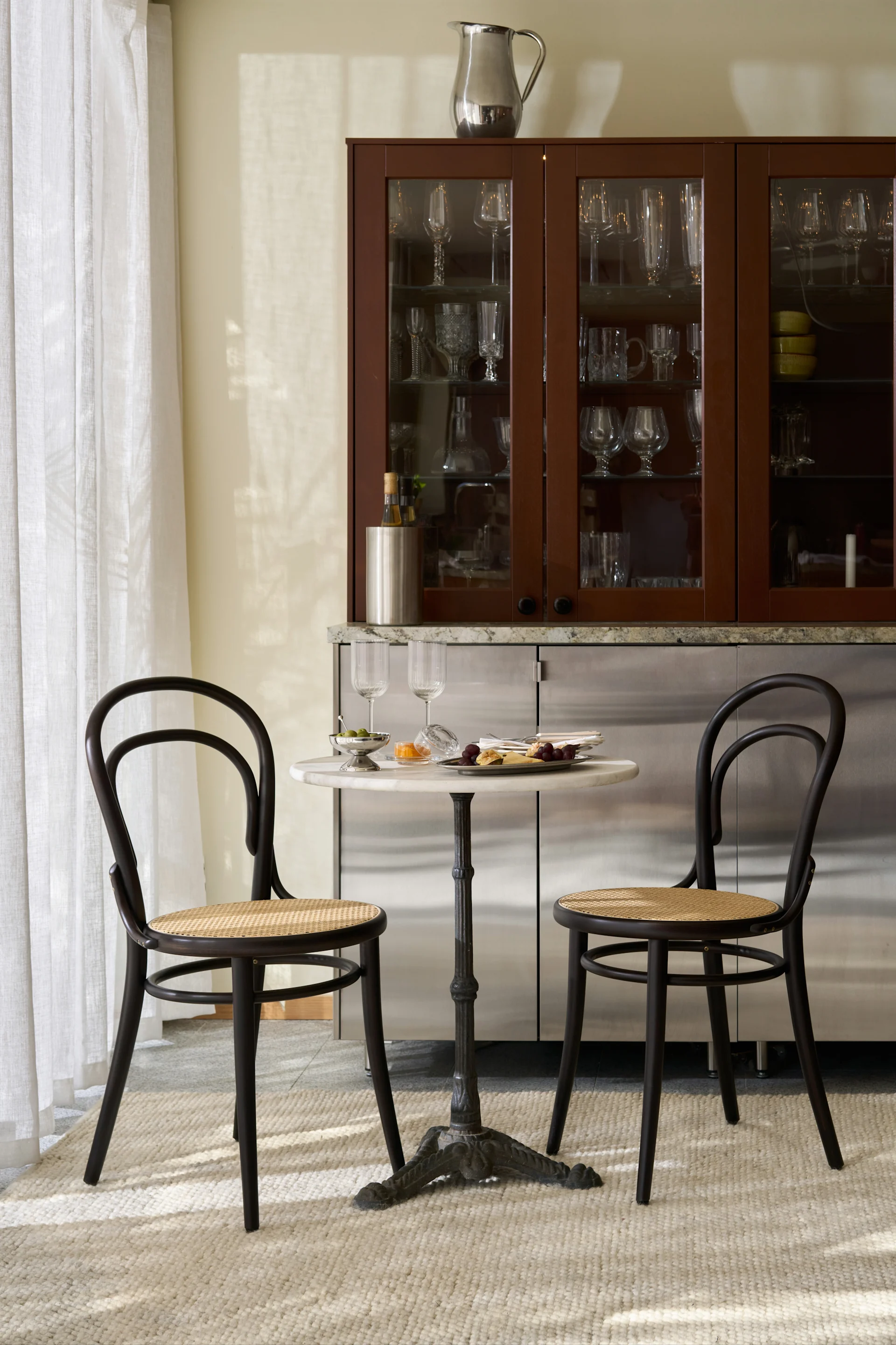 En stilfull matplats med ett bistrobord i marmor, två mörka trästolar och ett barskåp fyllt med glas.