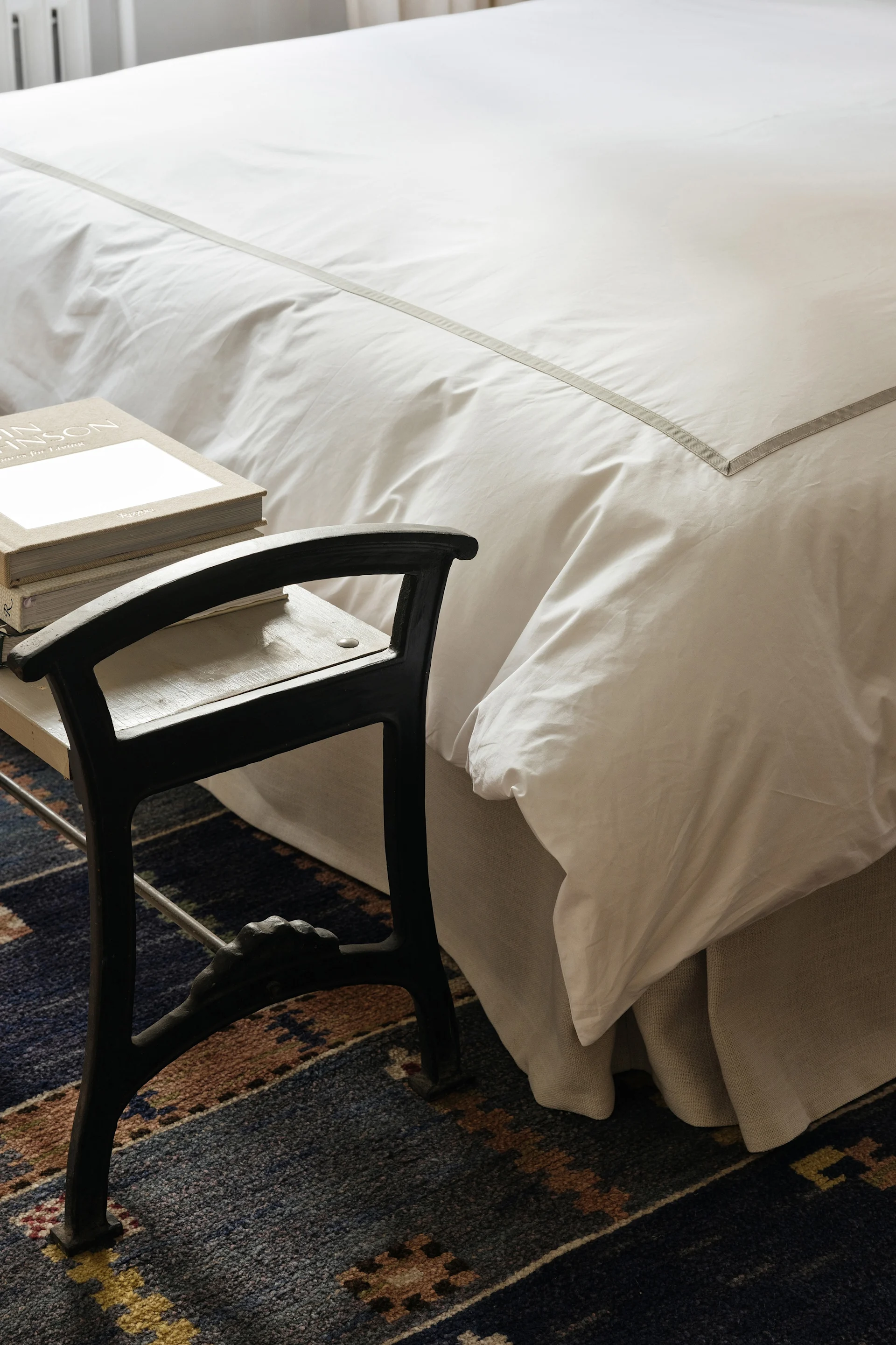 En säng med vita sängkläder och en grå rand bredvid en mörk pall med böcker på en mönstrad blå matta.