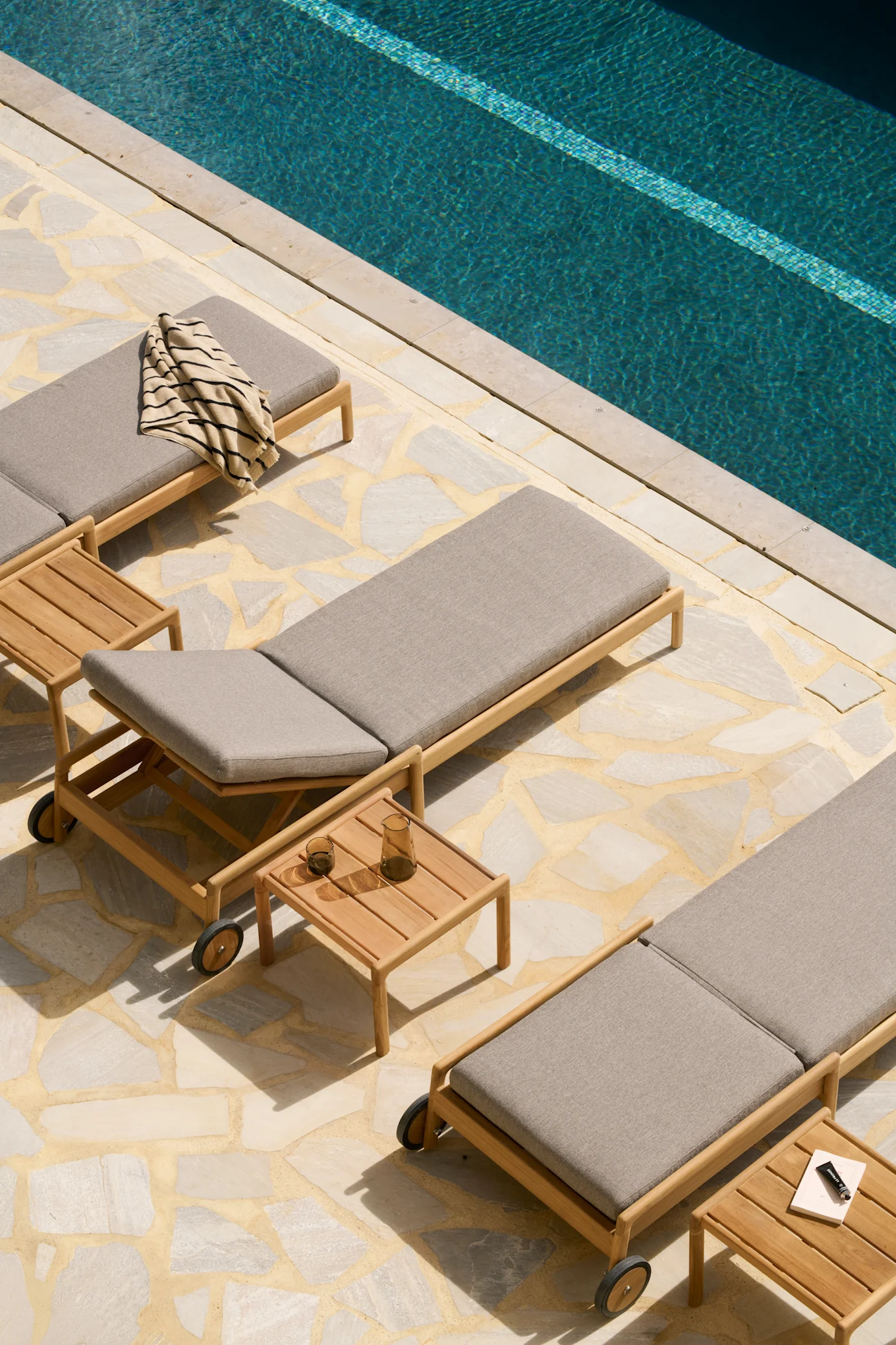 Överblick av tre träsolsängar med grå dynor, två sidobord och en randig handduk vid en blå pool på en stenlagd uteplats.