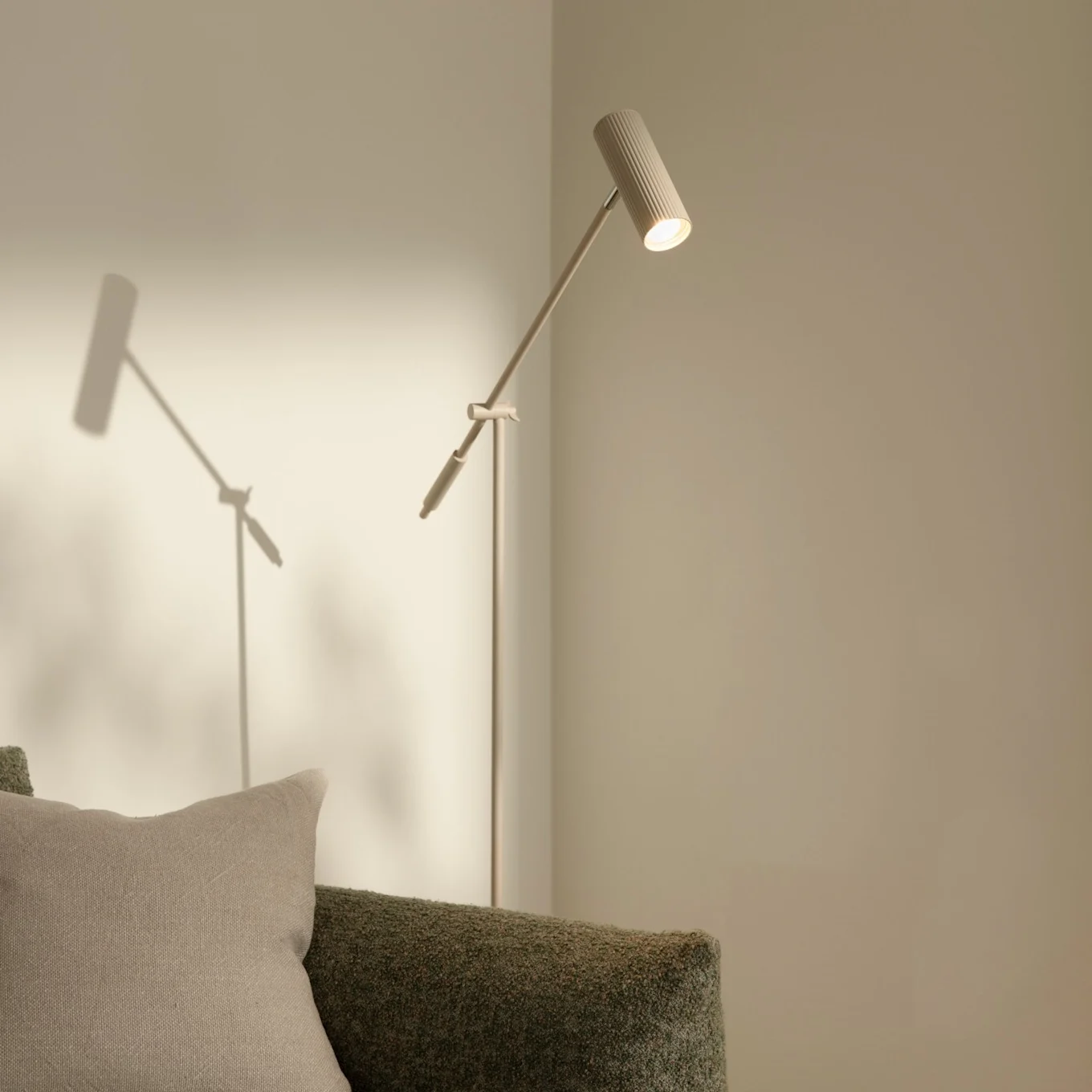 En modern, smal golvlampa med räfflat huvud som lyser upp en ljus vägg, bredvid en grön texturerad soffa med en beige kudde.