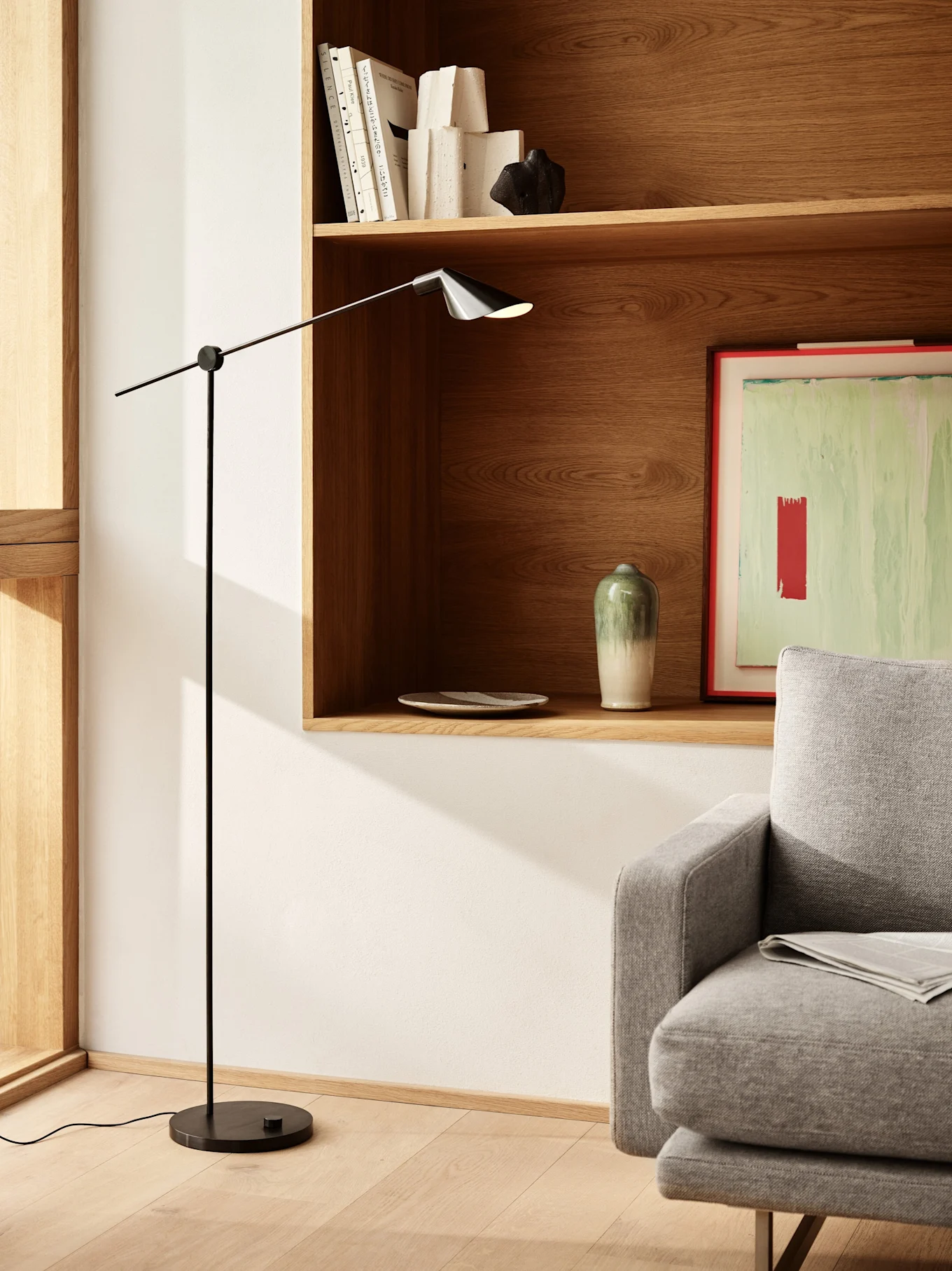 Ett modernt vardagsrum med en elegant svart golvlampa, trähyllor med böcker och inredning, samt en del av en grå soffa.