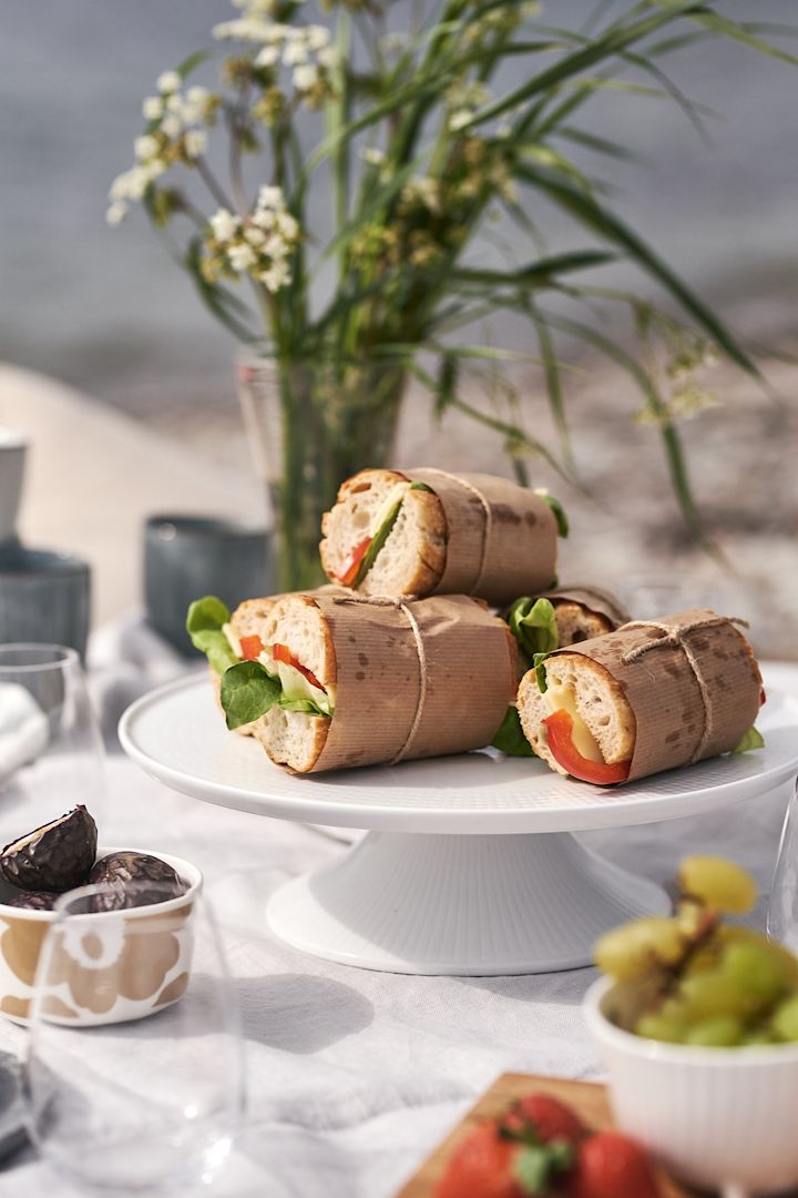 Picknick på stranden med mackor inslagna i vaxat papper på ett vitt tårtfat. 