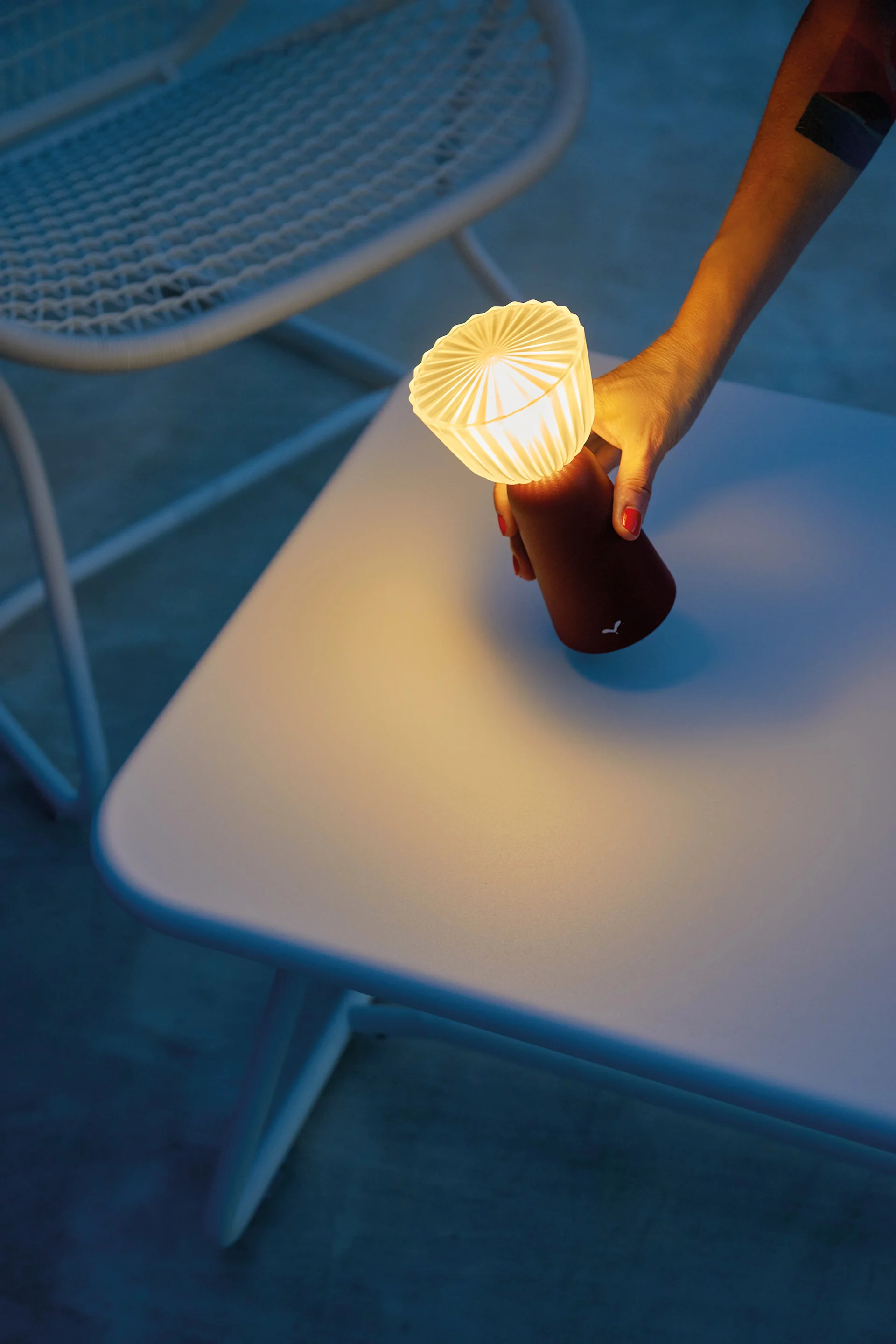 Hand med rött nagellack håller en liten glödande bärbar lampa med veckad topp över ett ljust bord. Inspiration till din balkong 2026.