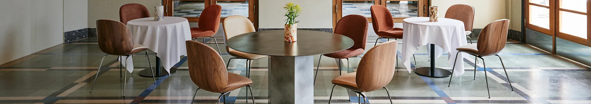 En elegant caféinteriör med ett stort centralt bord, två mindre bord med vita dukar och olika stolar på ett mönstrat golv.