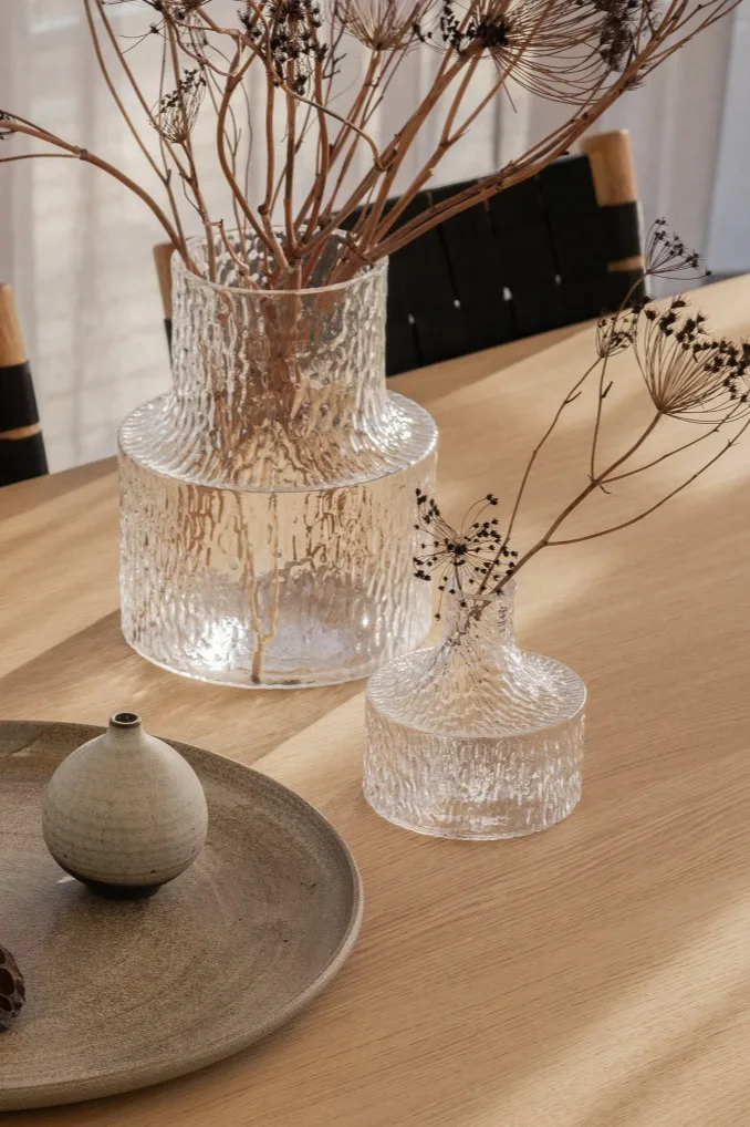 Två texturerade klara glasvaser med torkade grenar, en liten keramisk kruka på en grå tallrik, allt på ett ljust träbord.