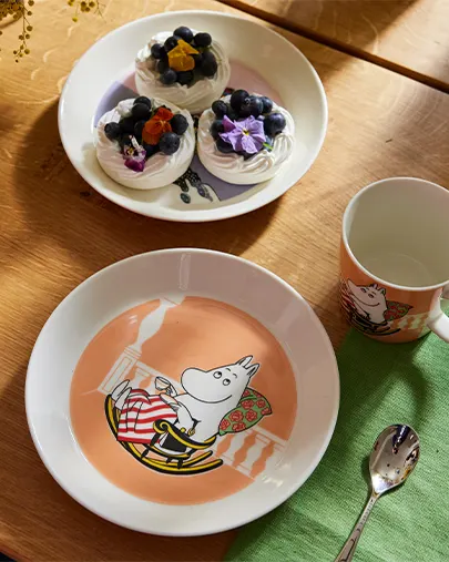 Mumin-tallrik, mugg och sked bredvid en tallrik med maränger med blåbär och blommor på ett träbord.