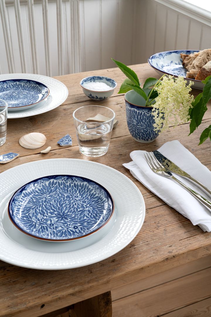 Tips på midsommardukning med blåvitt porslin och vit linneservett ledigt vikt vid sidan om, med besticken vilade ovanpå. 