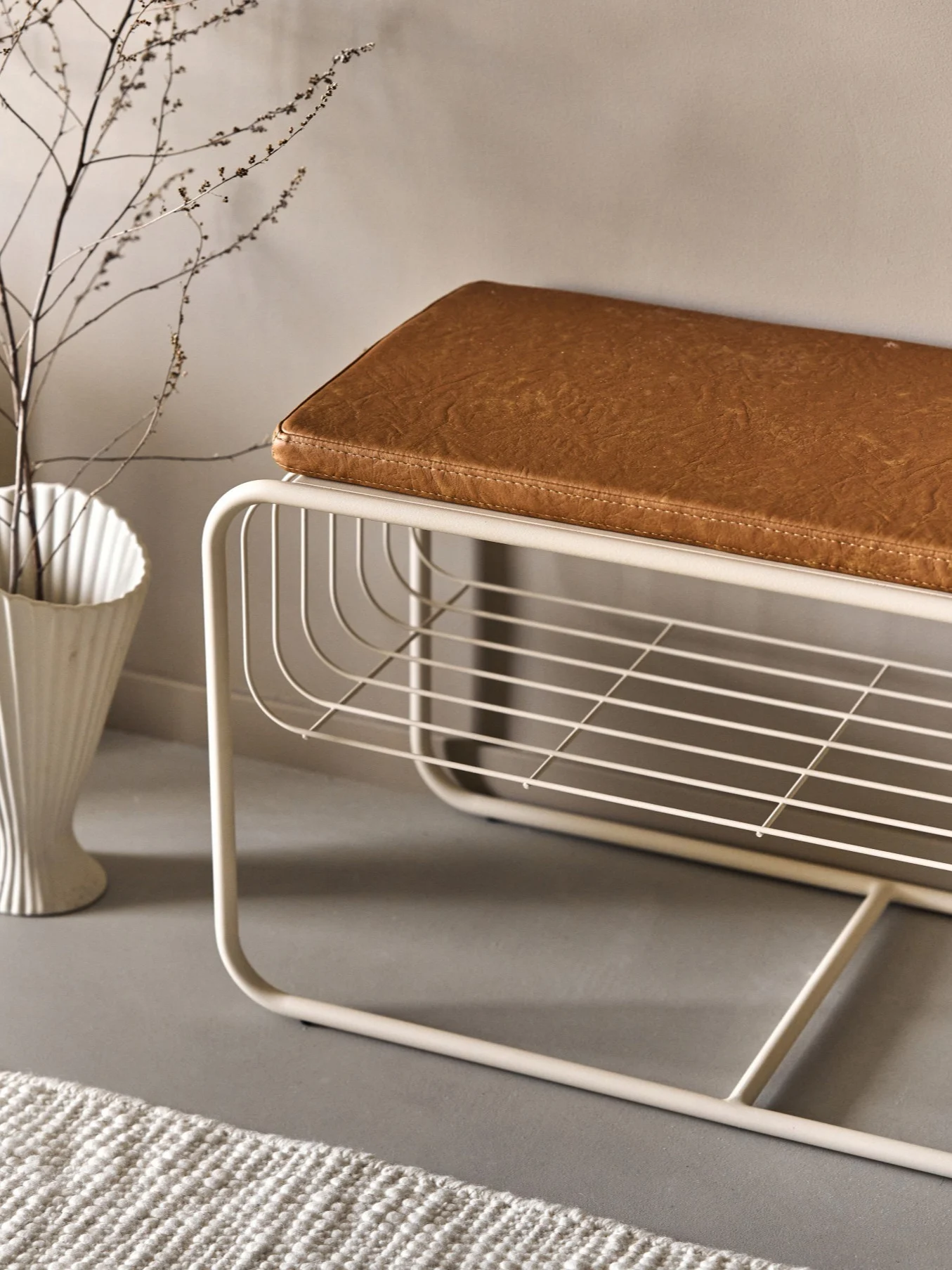 Modern beige metallbänk med brun konstläderkudde och öppen spalthylla. En vit vas med grenar och en matta finns i närheten.