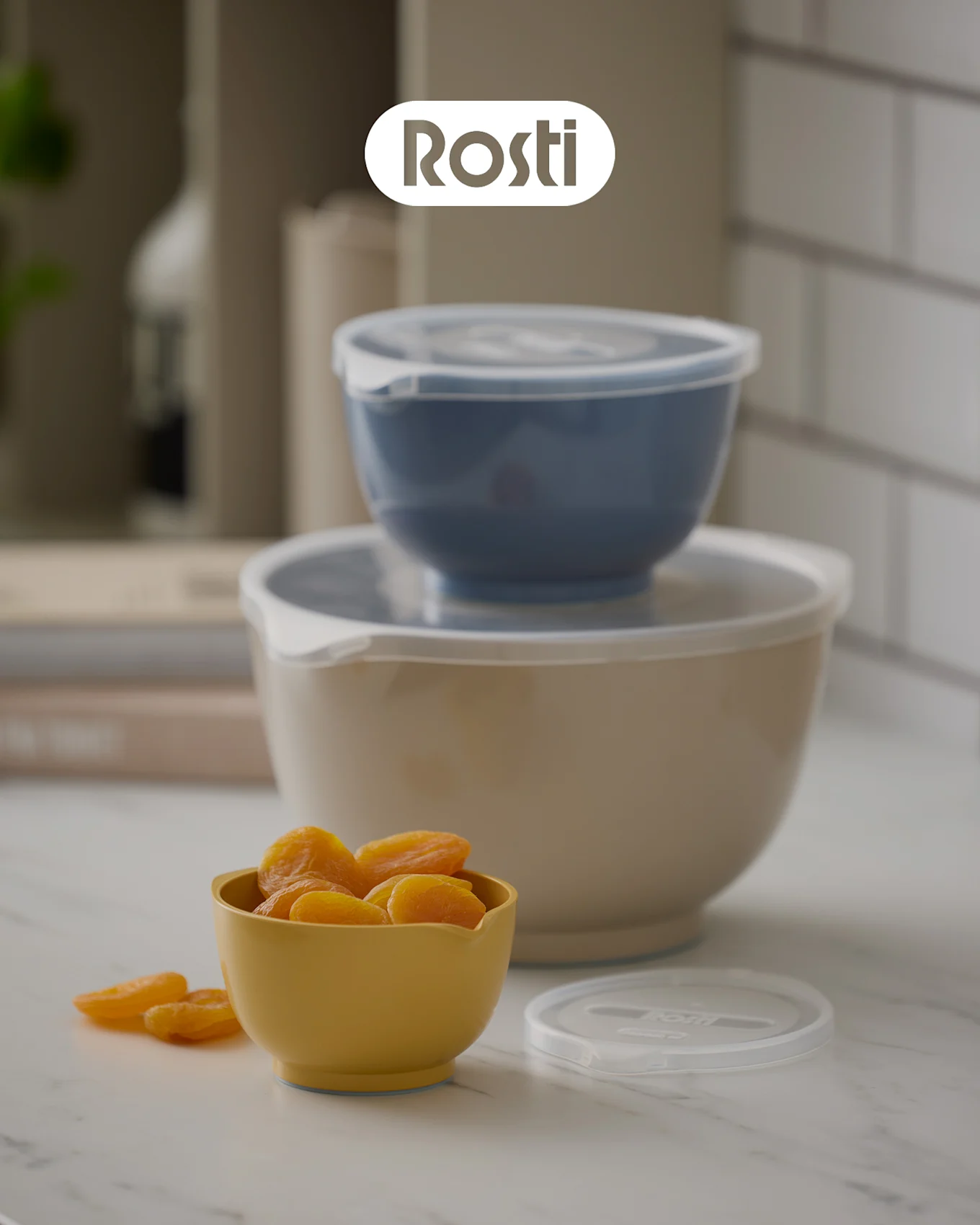 Tre Rosti-skålar på en bänk: en beige skål med en blå staplad ovanpå, båda med lock, och en liten gul skål fylld med torkade aprikoser.
