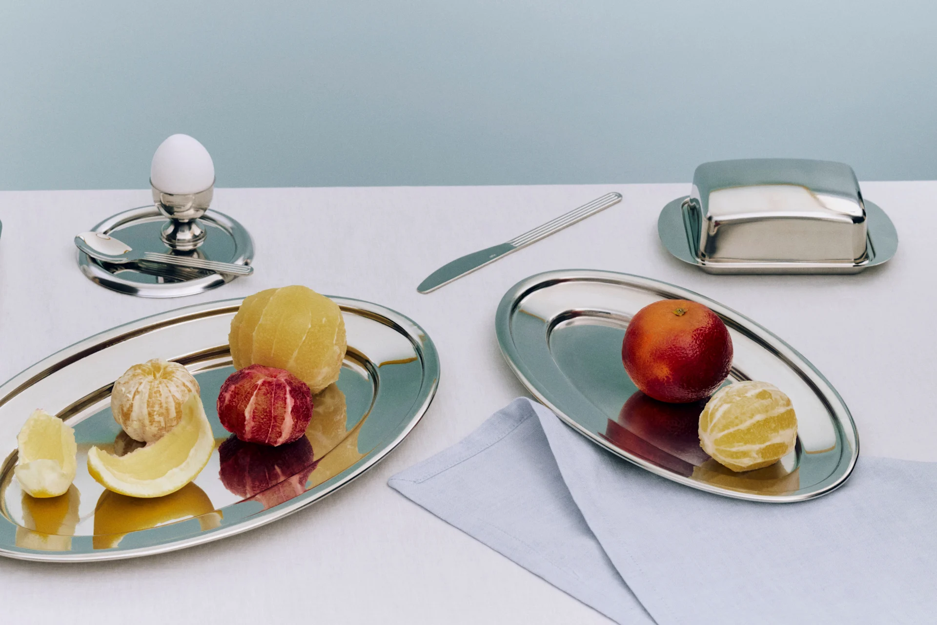 Frukostdukning med silverbrickor med skivat bröd, ett kokt ägg, skalade citrusfrukter, en blodapelsin, en smörbytta och en kniv.