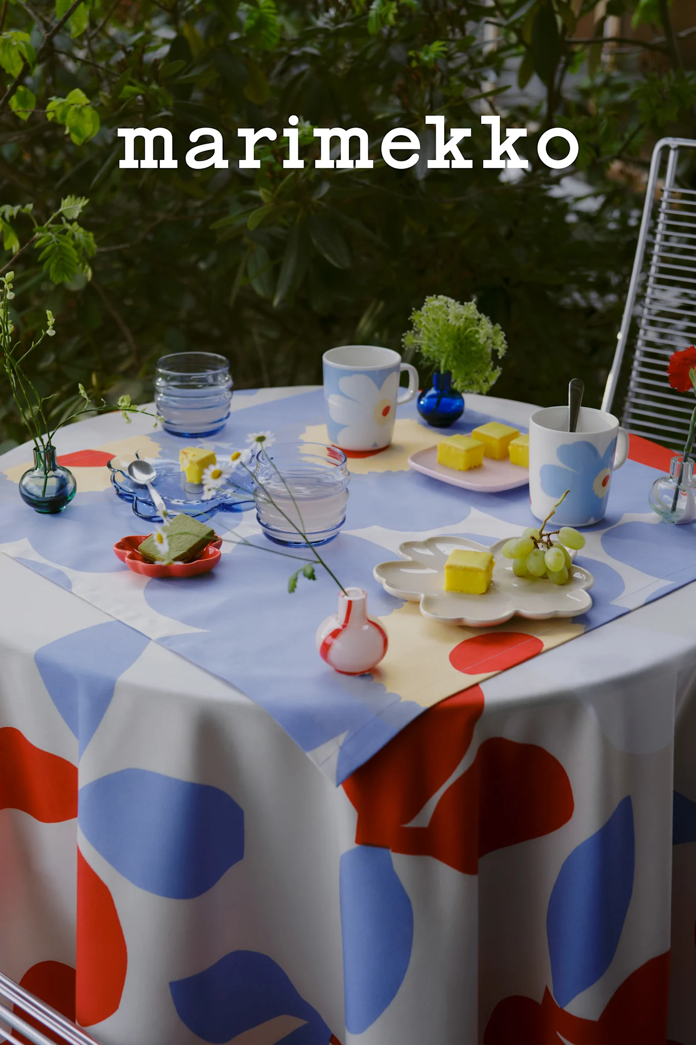 Marimekko-logotyp. Ett utomhusbord dukat med en blå, röd och vit abstrakt mönstrad duk, blommiga muggar, gula kakor, vindruvor och små vaser.