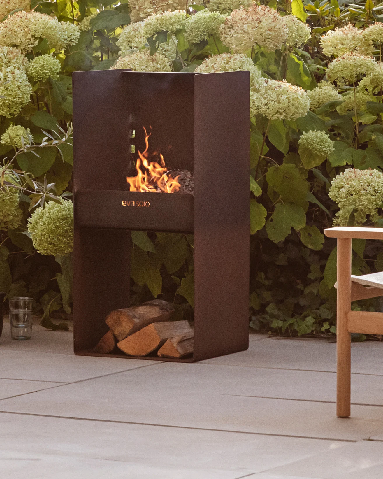 Modern mörk metall utomhuseldstad med brinnande eld, staplade vedträn undertill, omgiven av gröna växter och en uteplats.