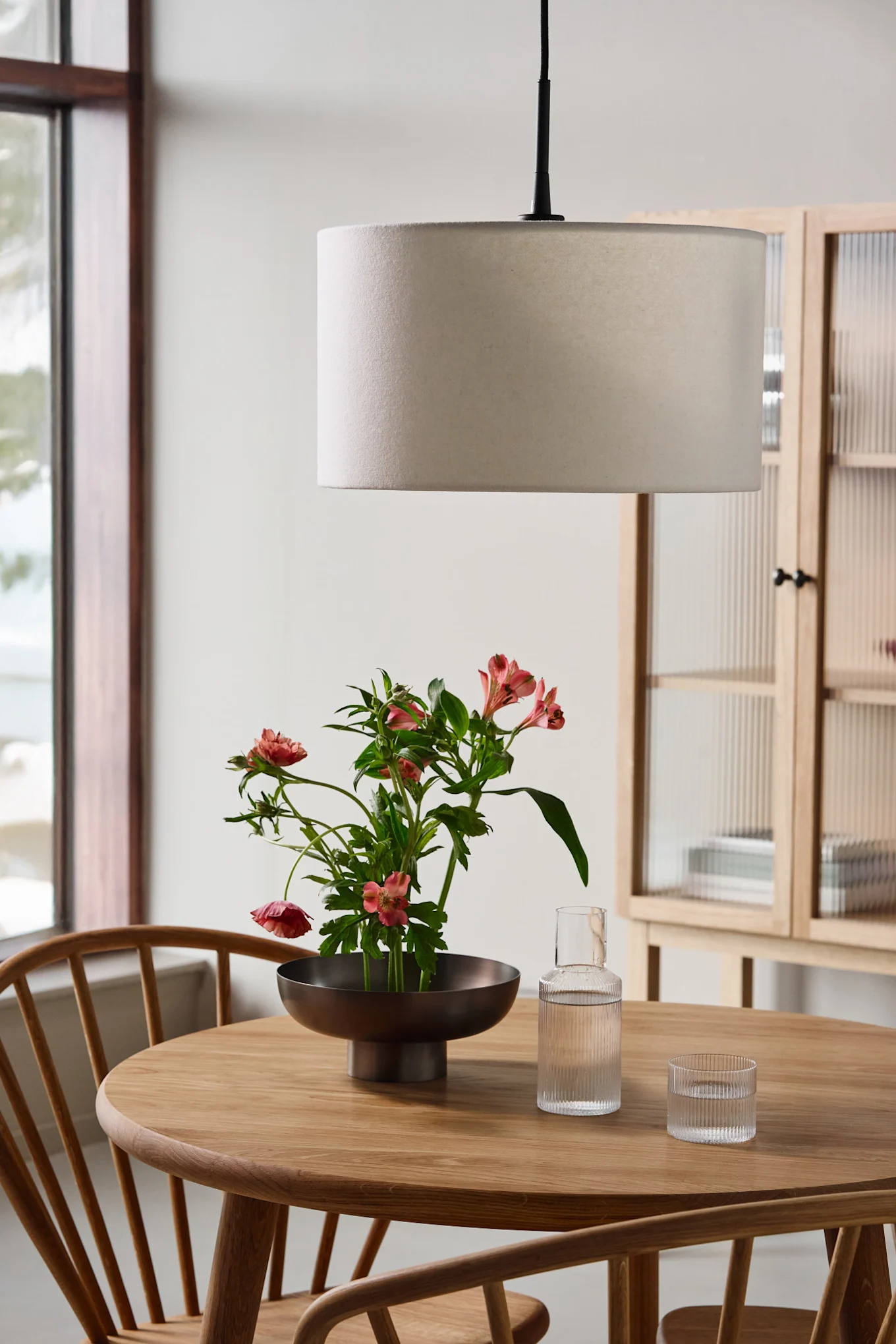 Ett runt träbord med en skål rosa blommor, en glaskaraff, ett glas och en trumformad taklampa.