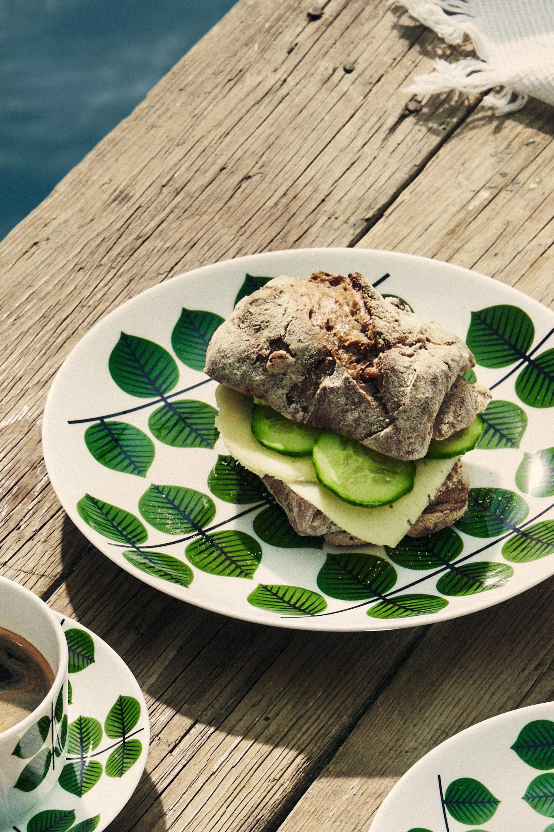 En ost- och gurksmörgås på en vit tallrik med gröna blad, bredvid en kaffekopp på en träbrygga, med blått vatten i bakgrunden.