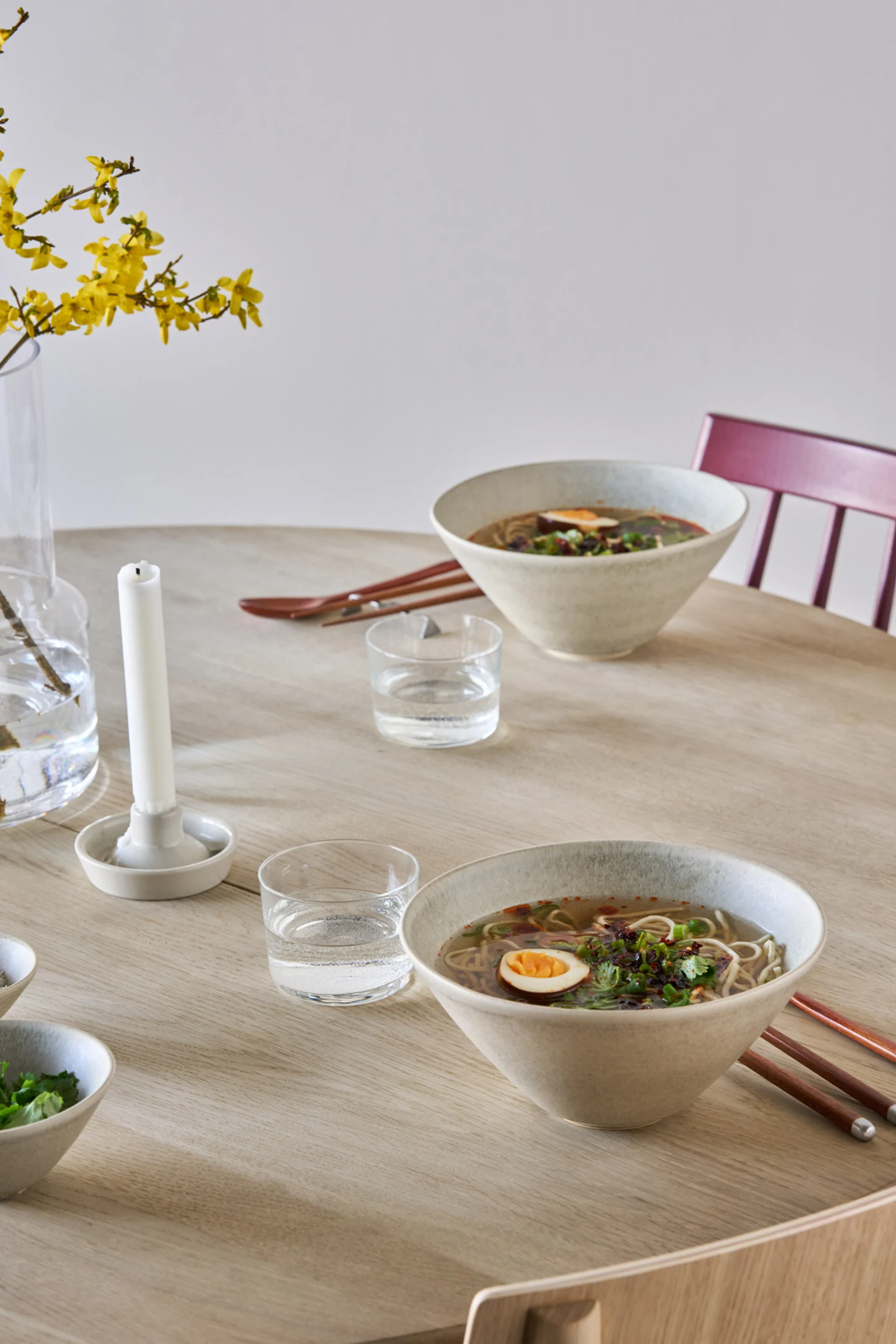 Två skålar ramen med ägg och garnering på ett ljust träbord, med vattenglas och gula blommor i en vas.