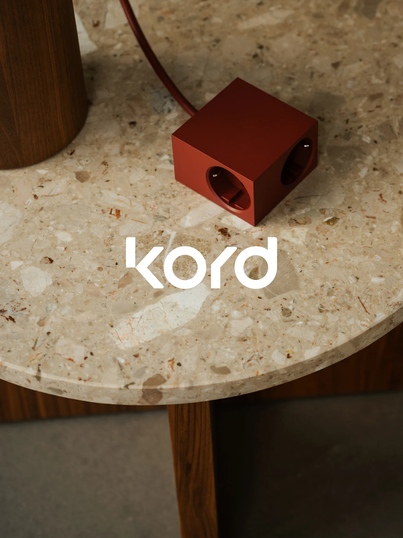 Ett mörkrött, kubformat grenuttag med två synliga uttag vilar på ett runt marmorbord, med den vita "kord"-logotypen.