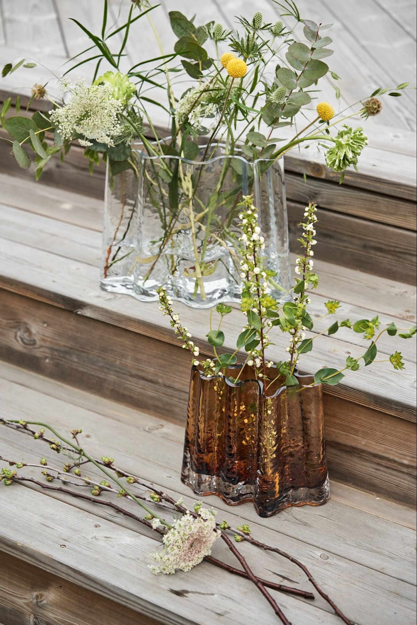 Gry wide vaser i färgerna clear och cognac står på ett altandäck i trä med vilda vårblommor i sig.