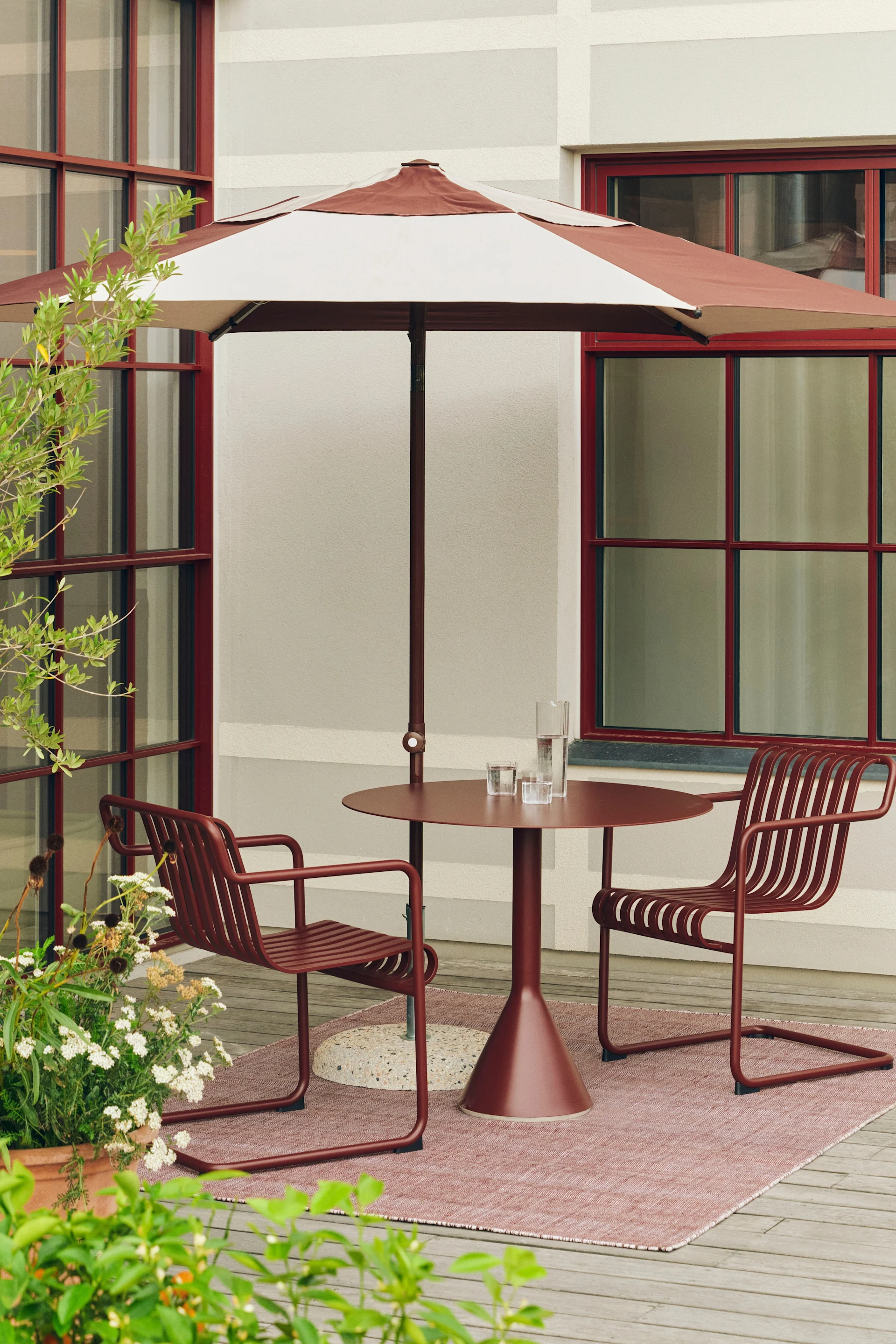 Uteplats med rödbrunt randigt parasoll, matchande runt bord och två ribbstolar på en rosa matta.