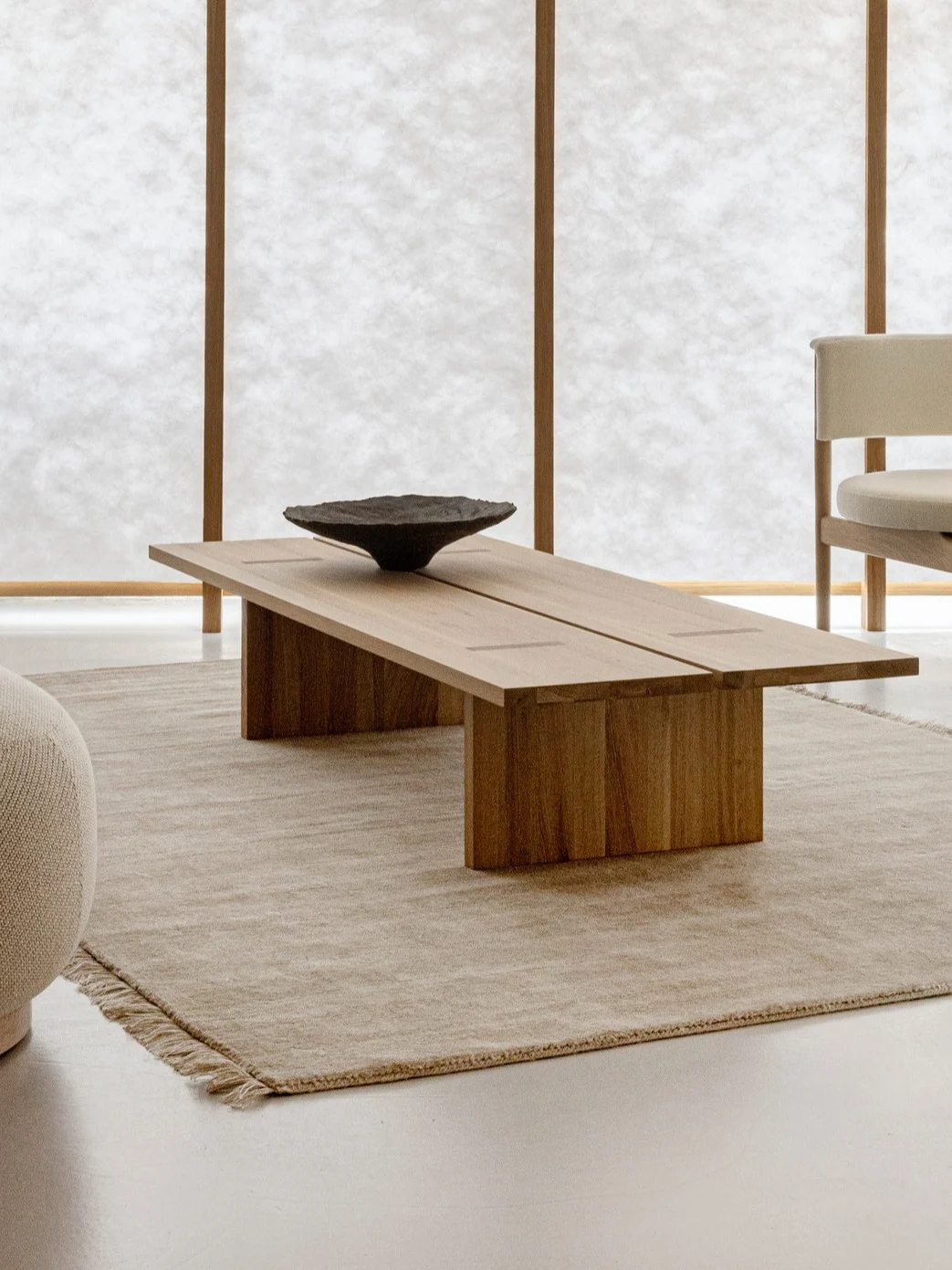 Minimalistiskt vardagsrum med soffbord i trä, beige matta, fåtölj och stol framför pappersskärmar.