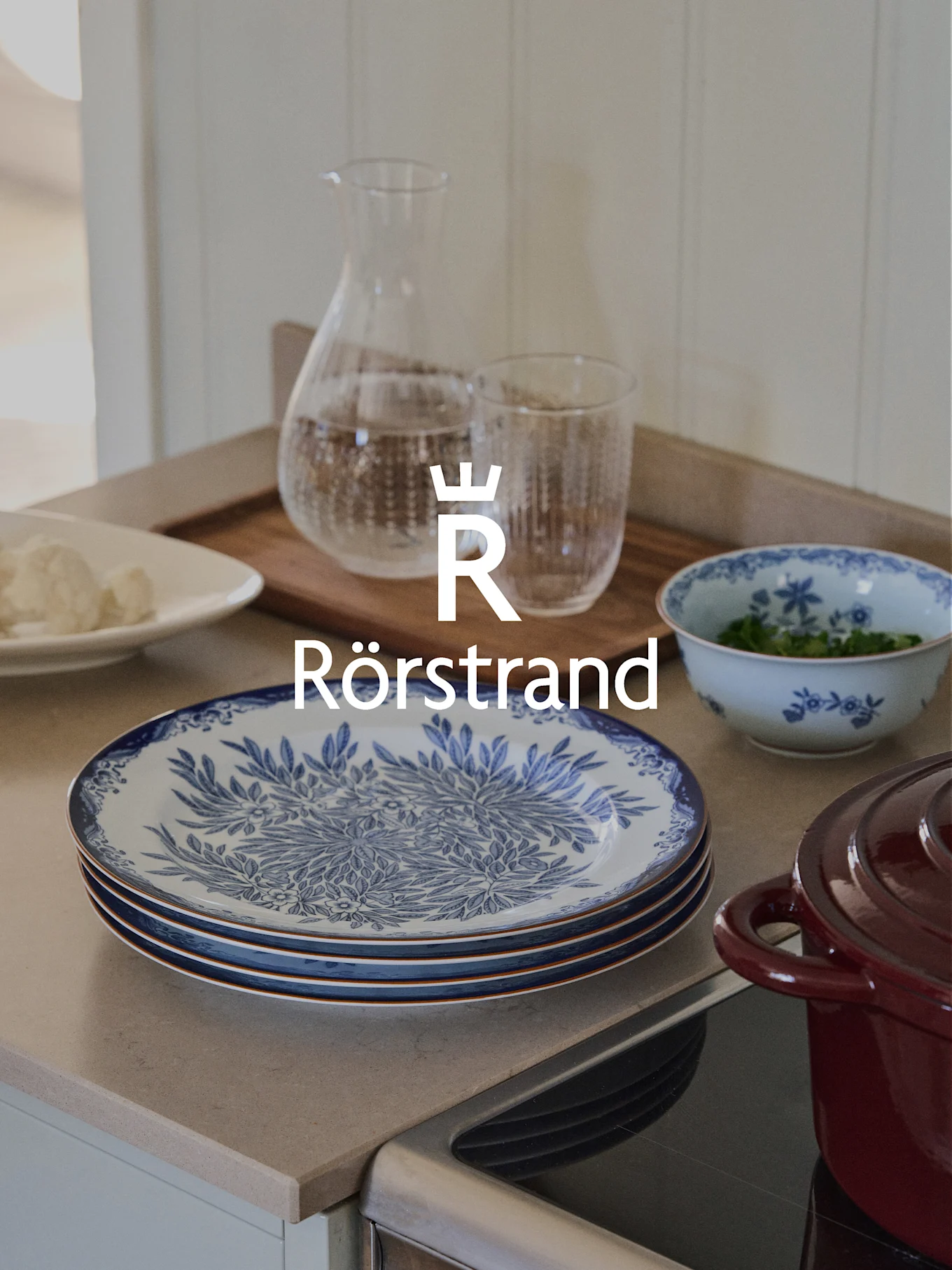 Rörstrand-logotyp över köksbänk med blå och vita tallrikar, karaff, glas och röd gryta.