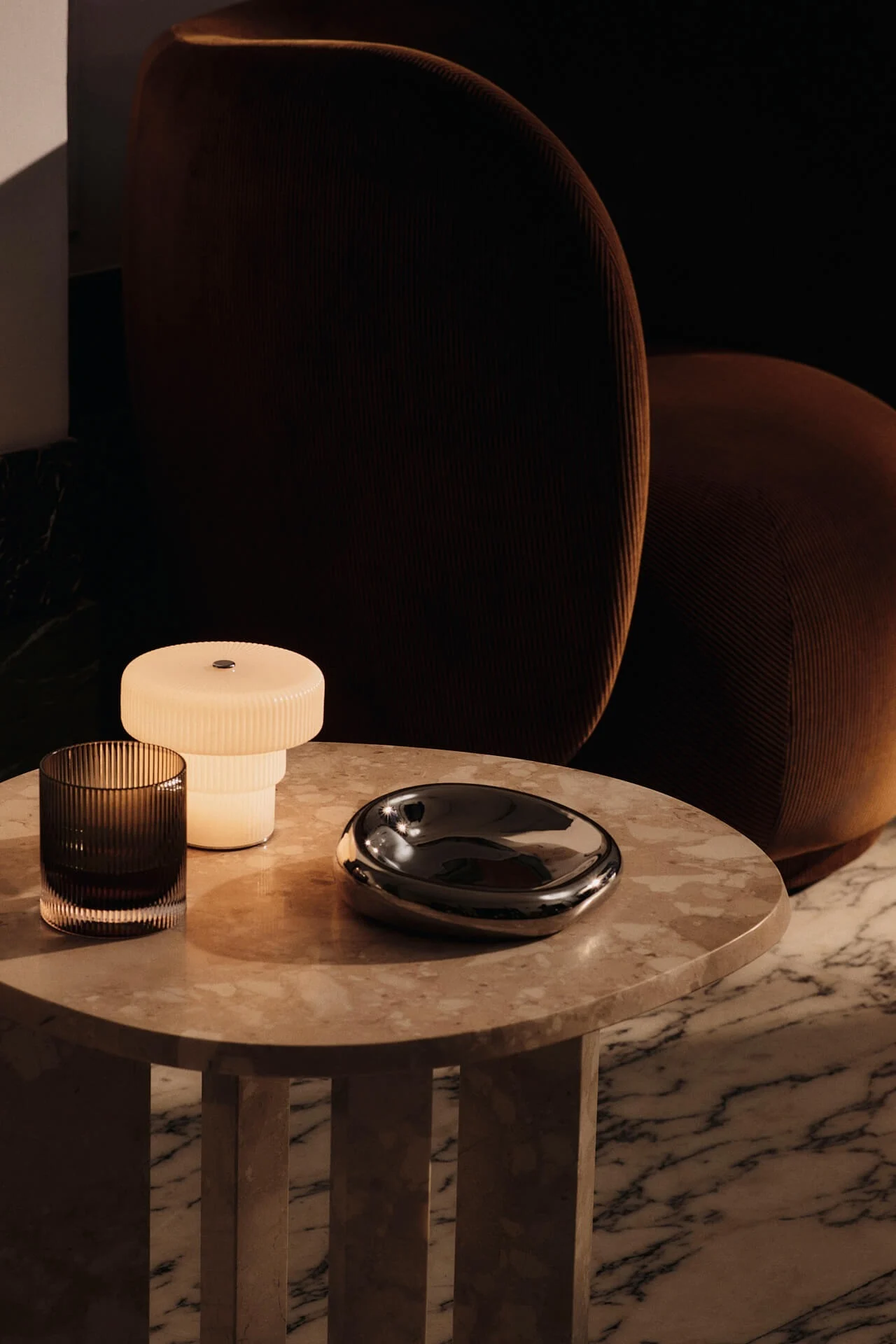 Ripple portabla lampa står på ett sidobord i sten med en Rico fotölj brevid.