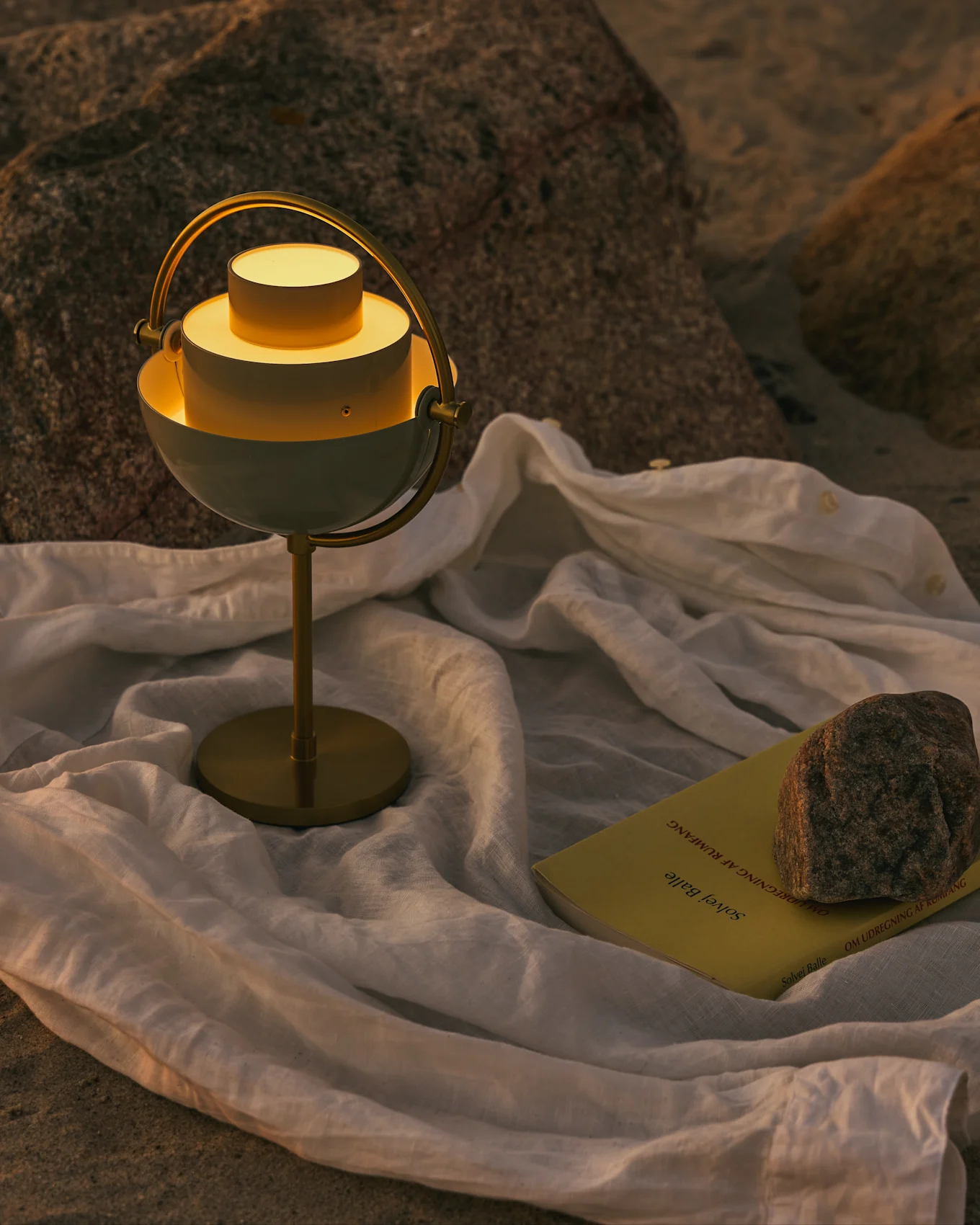 En modern vit och mässing bordslampa med varmt ljus, en gul bok och en sten på ett vitt tyg på en stenig yta.