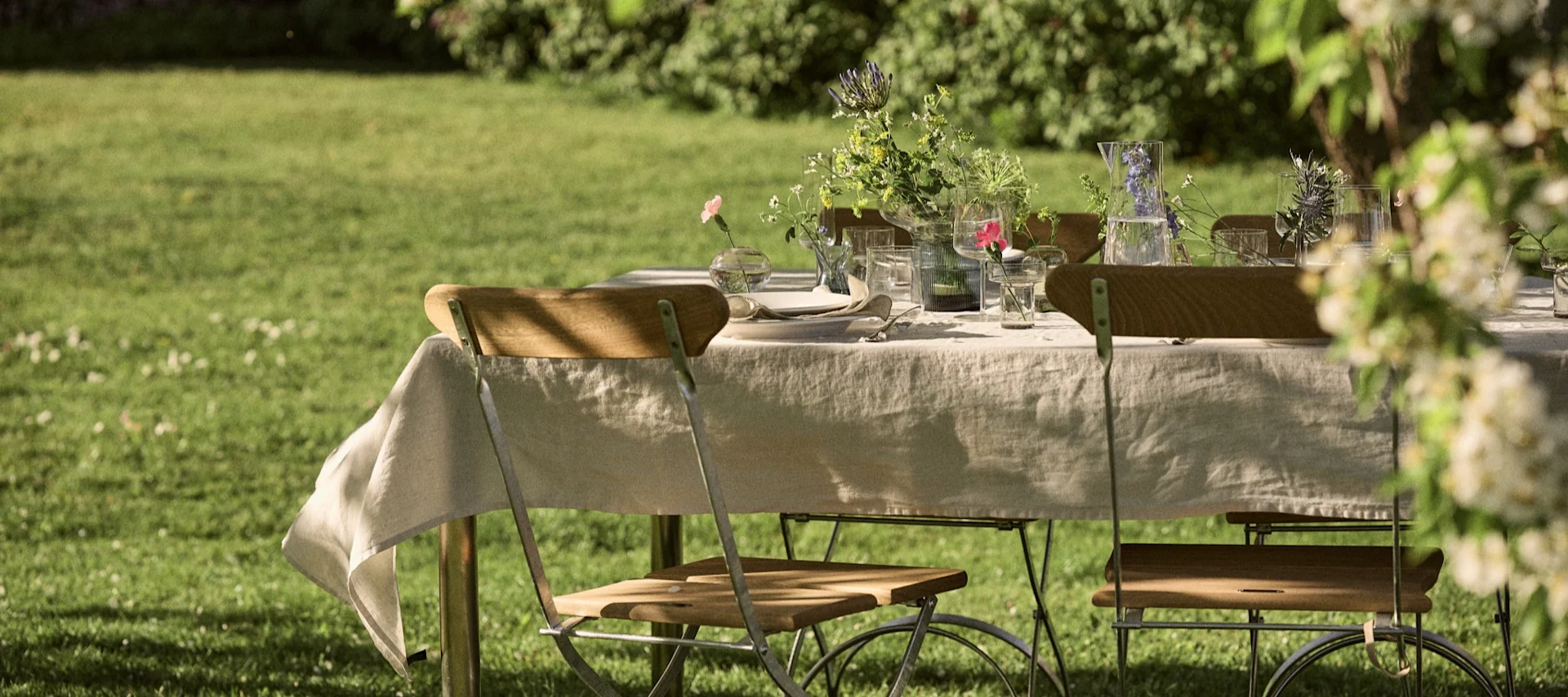 Ett utomhus matbord dukat med linneduk, trästolar, glas, tallrikar och vaser med vildblommor på en grön gräsmatta.