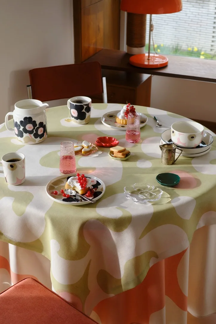 Ett runt bord med en grön, rosa och vit blommig duk, dukat med svart-rosa mönstrade muggar, tallrikar, en kanna, bakverk toppade med röda vinbär och rosa glas.
