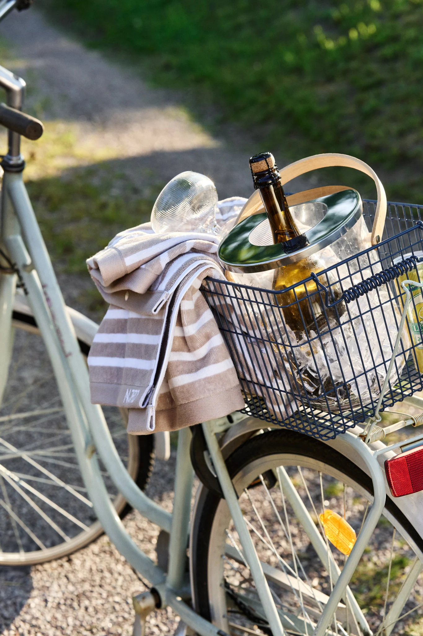 En cykelkorg innehåller en ishink med en kylande flaska och en randig handduk en solig dag.