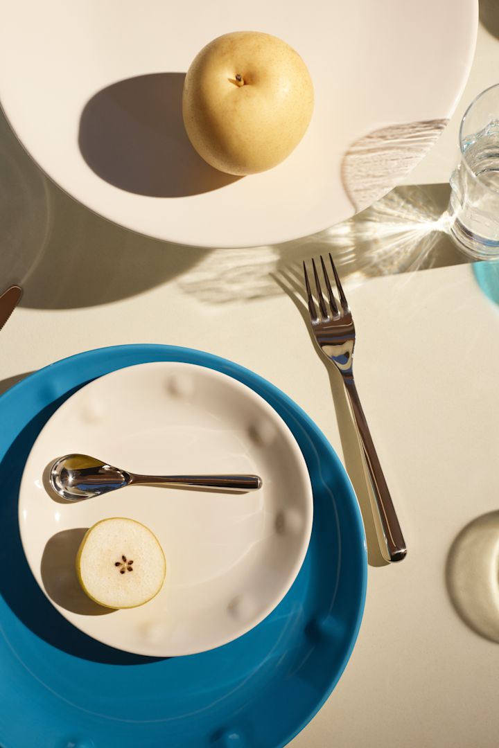 En vit skål placerad ovanpå en blå tallrik med ett glas och bestick från Solare Collection från Iittala. 