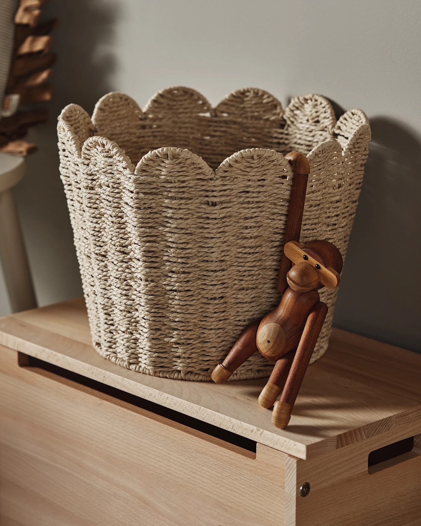 En ljus flätad korg med en vågig kant står på en träyta, med en träapafigur hängande från sidan.