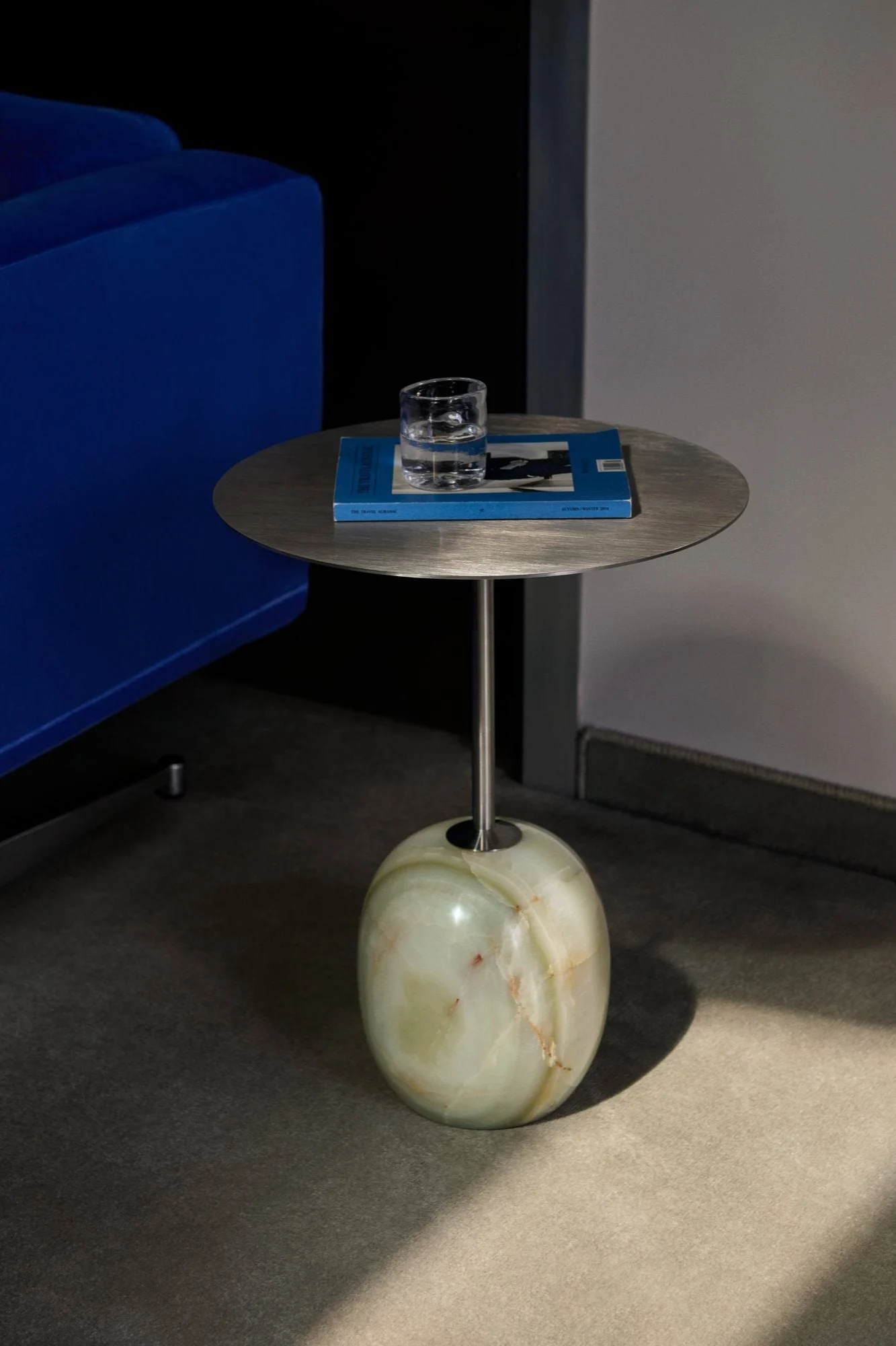 Sidobord med borstad metallskiva, rund grön onyxbas, vattenglas, blå bok, bredvid en blå soffa.
