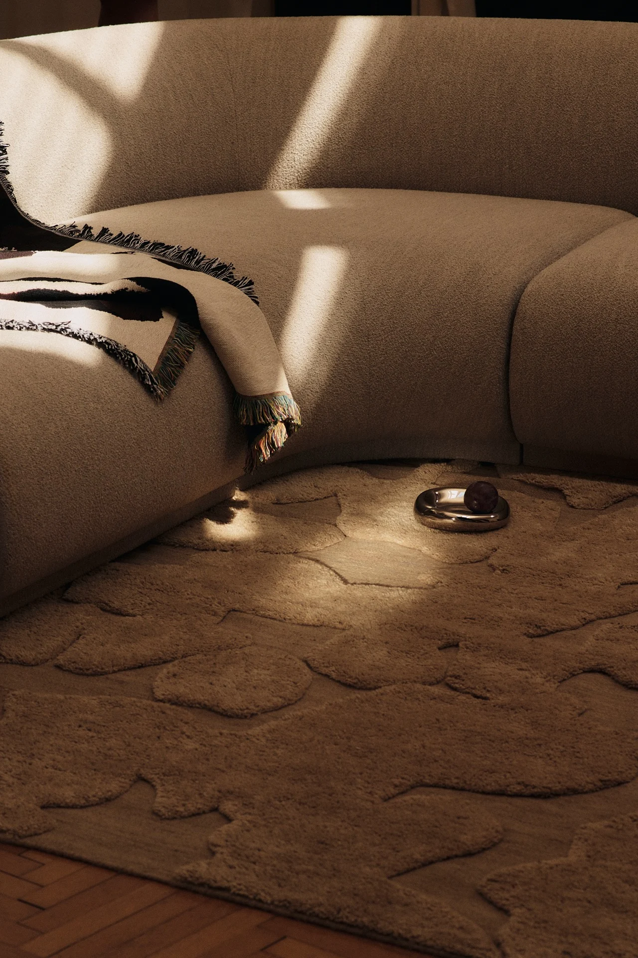 Svängd beige soffa med mönstrad pläd, organisk formad beige matta och en silverskål med mörk frukt i ett soligt rum.