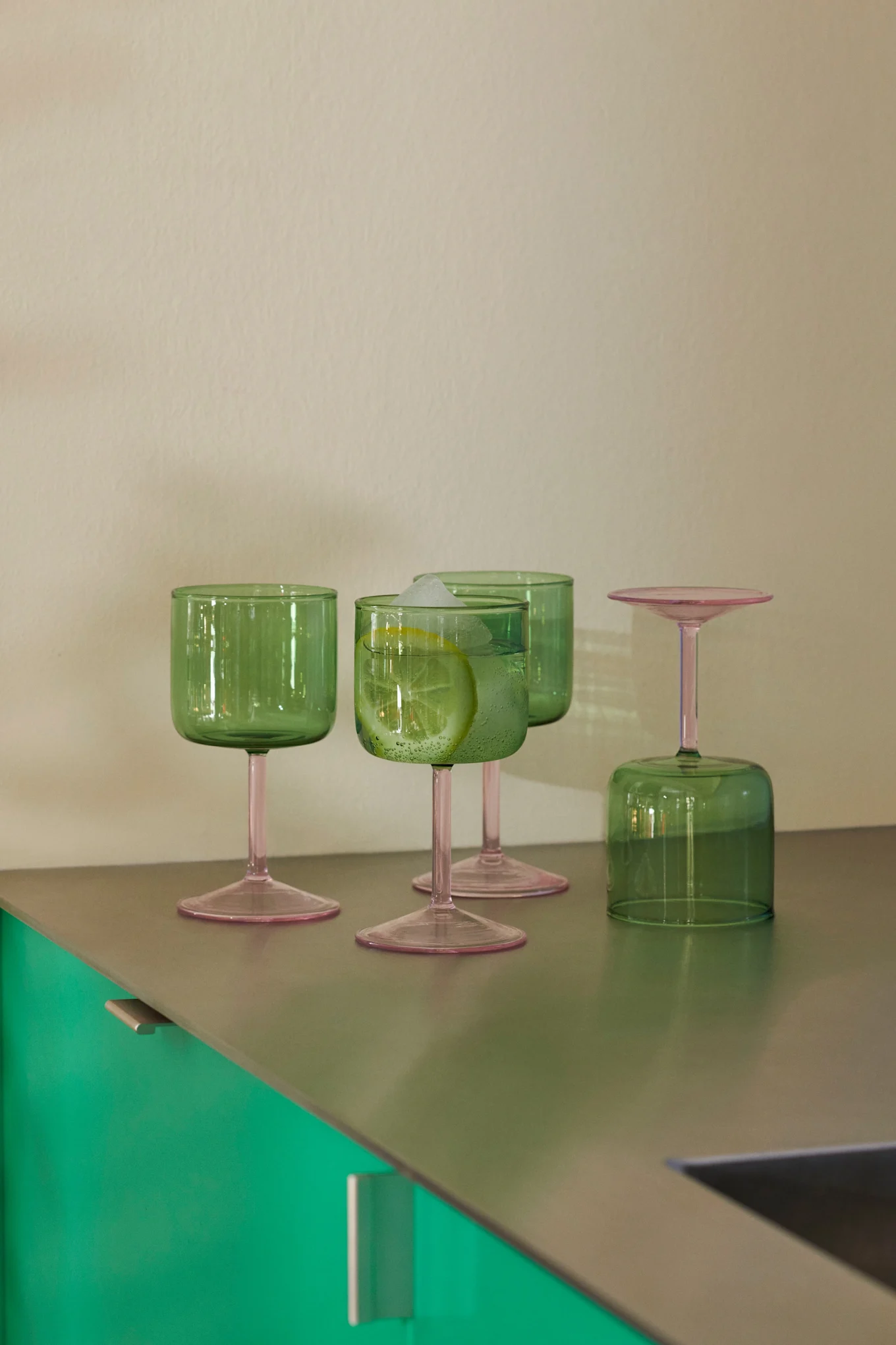 Fyra grönfärgade glas med rosa stjälkar på en metallisk bänkskiva. Ett glas innehåller en dryck med citron och is; ett annat är upp och ner.