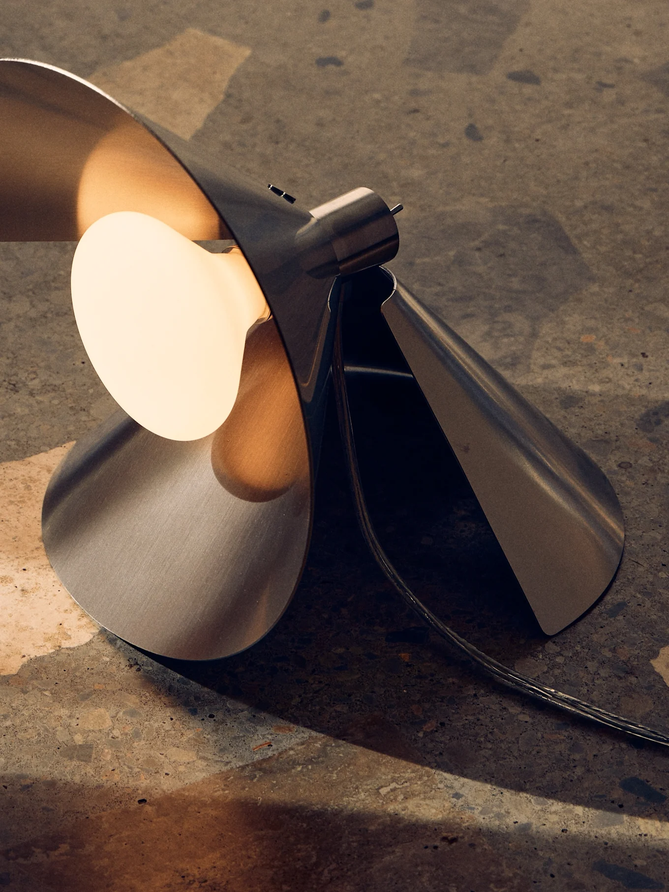 Modern borstad metallbordslampa med skulptural, vikt design och en varm, exponerad glödlampa på en texturerad yta.