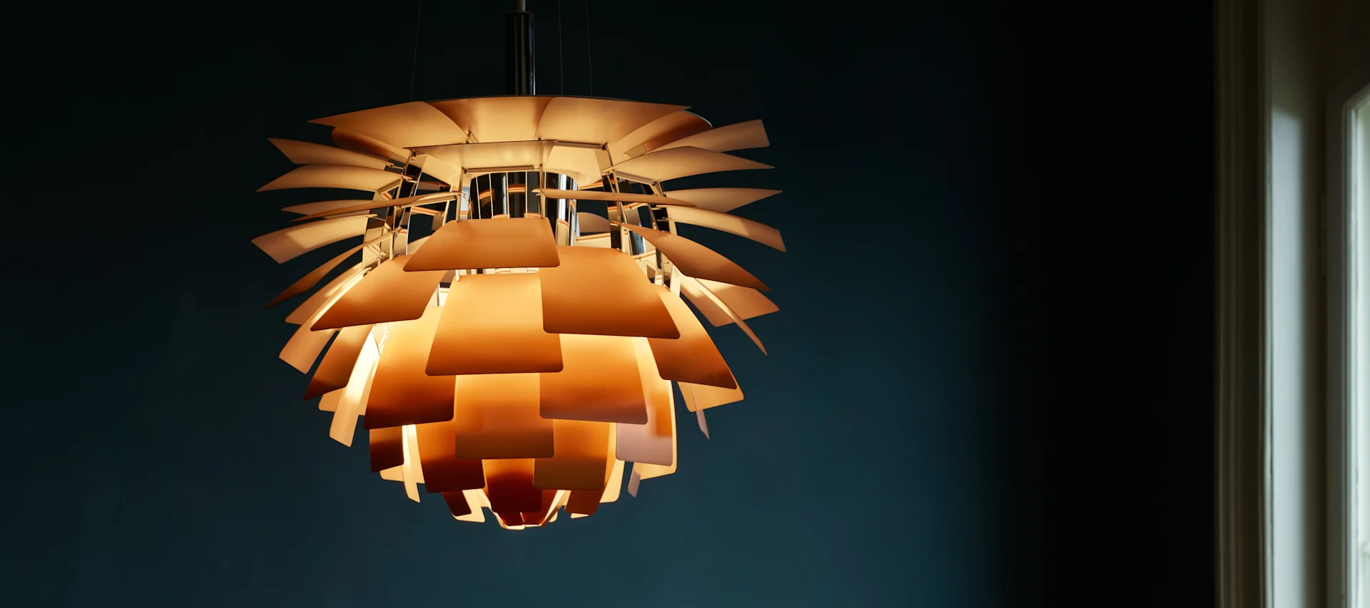 En lysande PH Artichoke pendel från Louis Poulsen hängande från ett tak med mörkblå bakgrund.