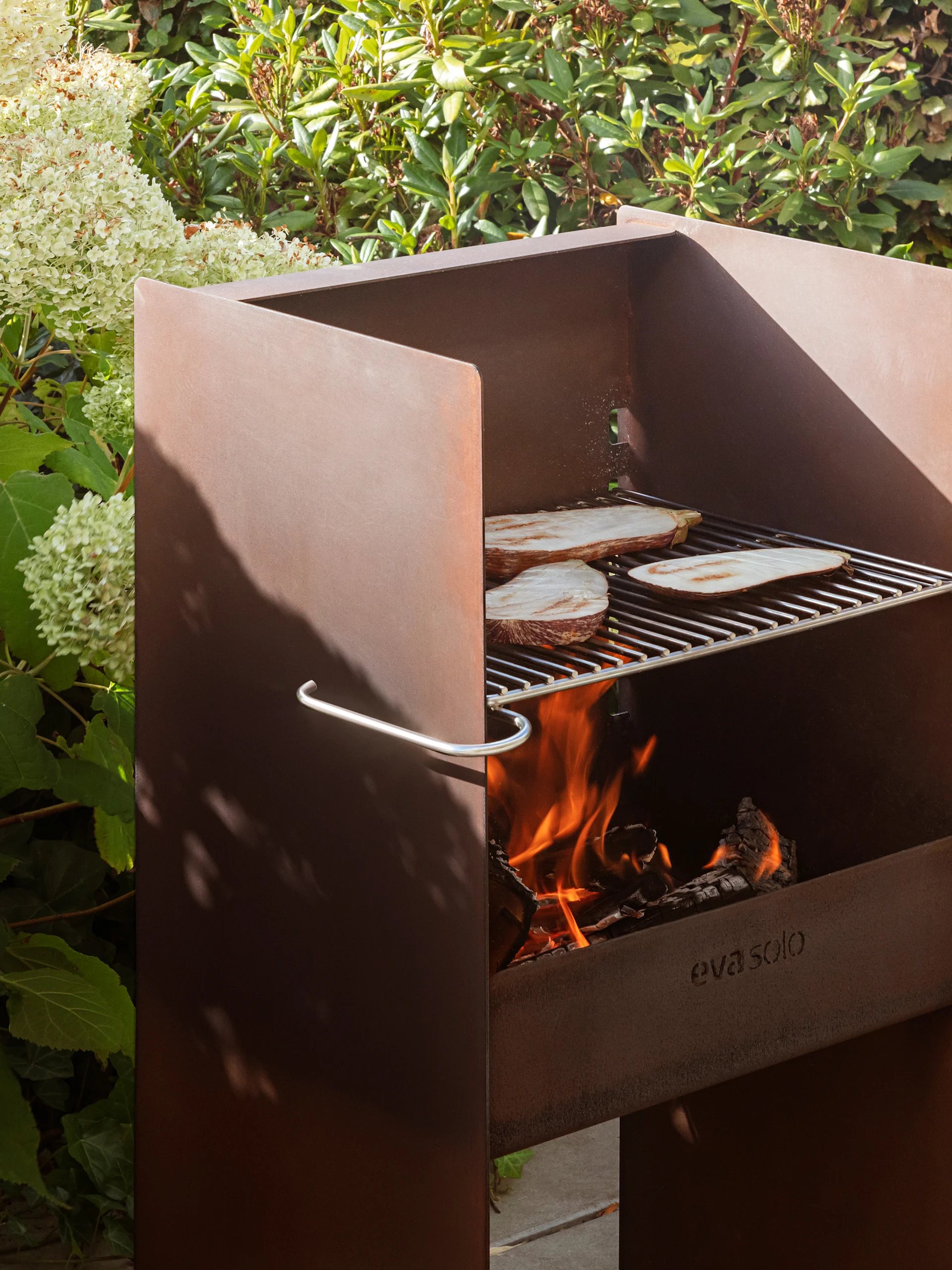Rostig metall utomhusgrill från Eva Solo med aubergine på gallret, brinnande eld och lummig grönska.