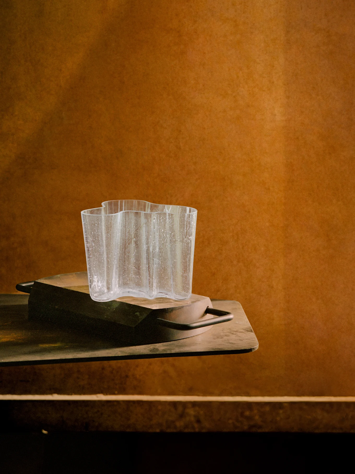 Klar, vågig glasvas med vattendroppar på en mörk träbricka, mot en varm brun texturerad bakgrund.
