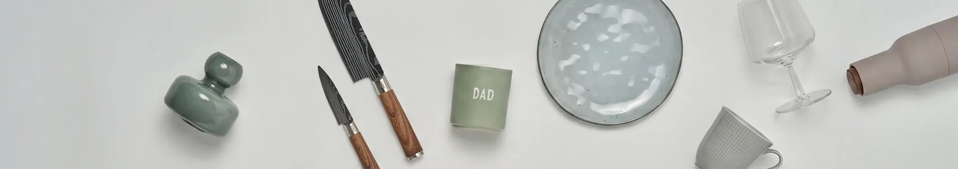 En flat lay av köks- och presentartiklar: en grön vas, två knivar, ett 'DAD'-ljus, en grå keramisk tallrik, ett vinglas, en beige flaska och en grå mugg.