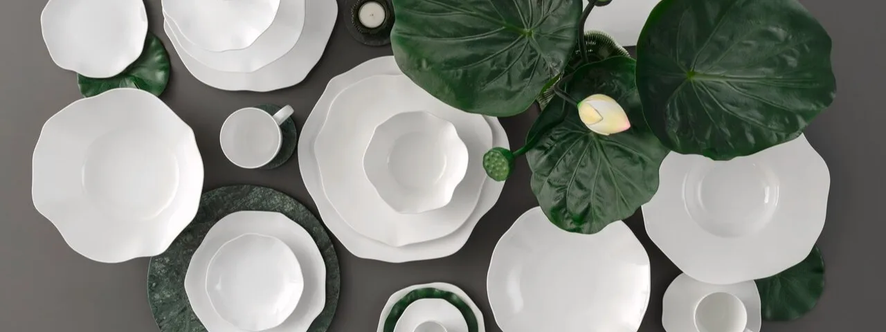 En överliggande bild av vit keramikservis med vågiga kanter, arrangerad med gröna blad och marmorunderlägg på en grå bakgrund.