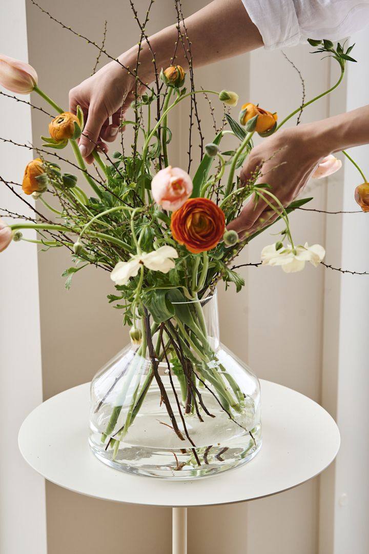Här ser du händer som arrangerar blommor i en klar trattformad vas som ingår i Solare-kollektionen från Iittala. 
