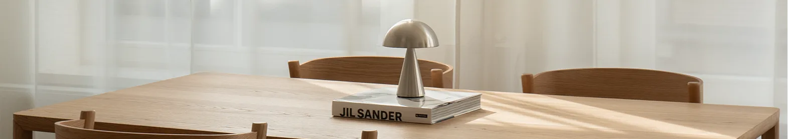 Ljust matbord i trä med fyra stolar. Silverfärgad svamplampa och bok på bordet. Solljus genom skira vita gardiner.
