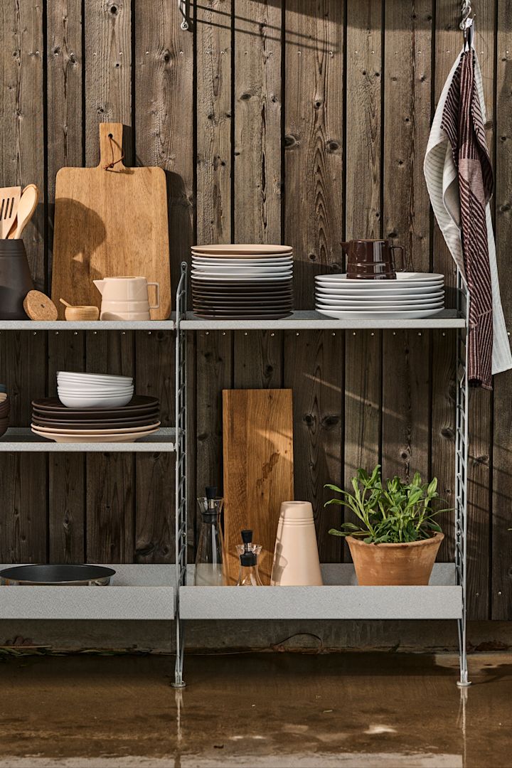 Bild som visar String Outdoor i galvaniserat stål, ett system som är anpassat för utomhusbruk och som bland annat kan användas till att bygga ett utekök, här fyllt med köksredskap, porslin och färska örter.