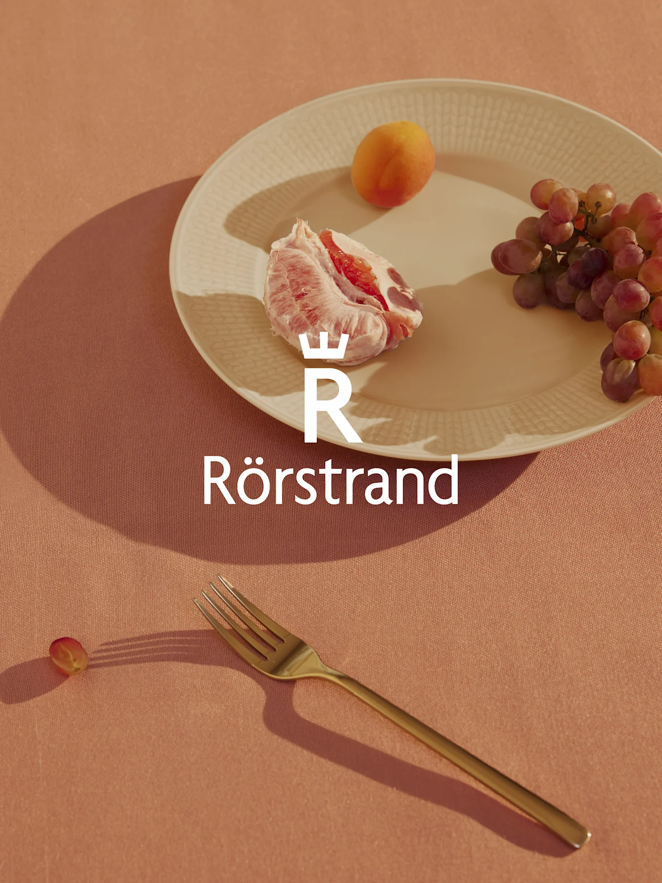 Rörstrand-logotyp över en tallrik med grapefrukt, aprikos och vindruvor, en gaffel och en ensam druva på en persikofärgad duk.