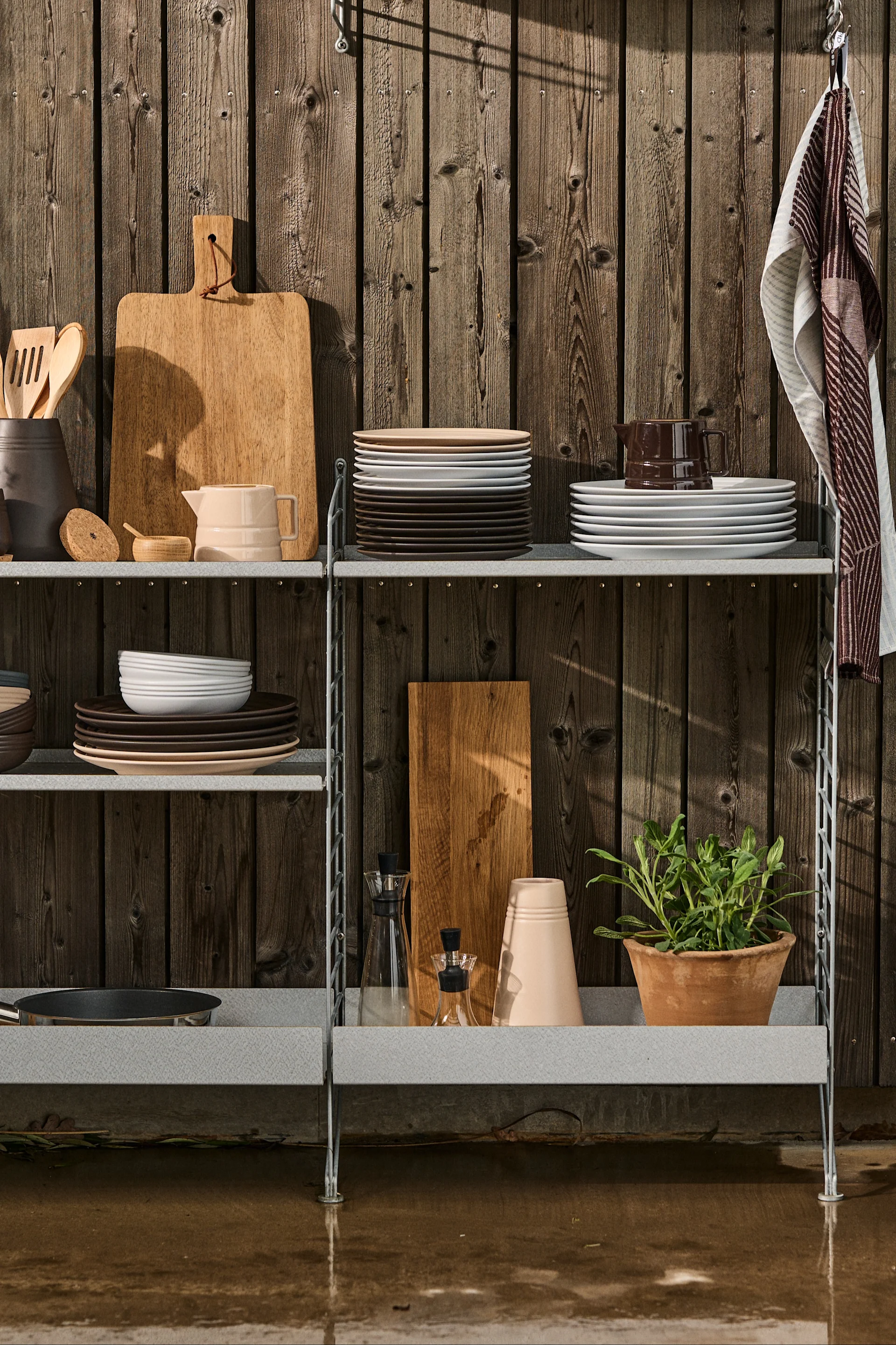 Bild som visar String Outdoor i galvaniserat stål, ett system som är anpassat för utomhusbruk och som bland annat kan användas till att bygga ett utekök, här fyllt med köksredskap, porslin och färska örter.